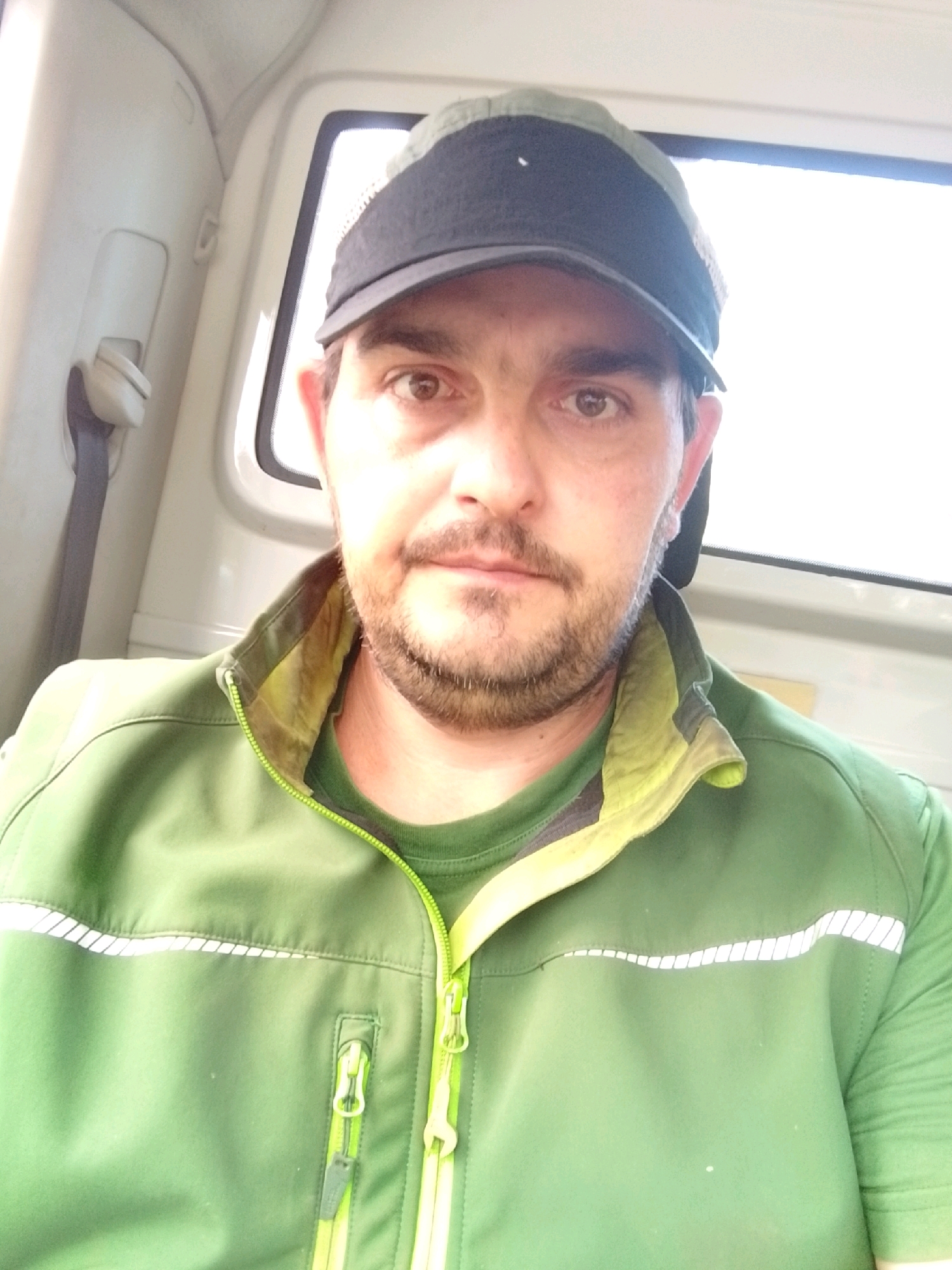 A man wearing a green jacket and a black cap is in the driver's seat of a vehicle. He is looking at the camera. The interior of the vehicle is visible in the background, including a seatbelt and a window. The scene appears to be a selfie taken inside a vehicle. The location is not specified.