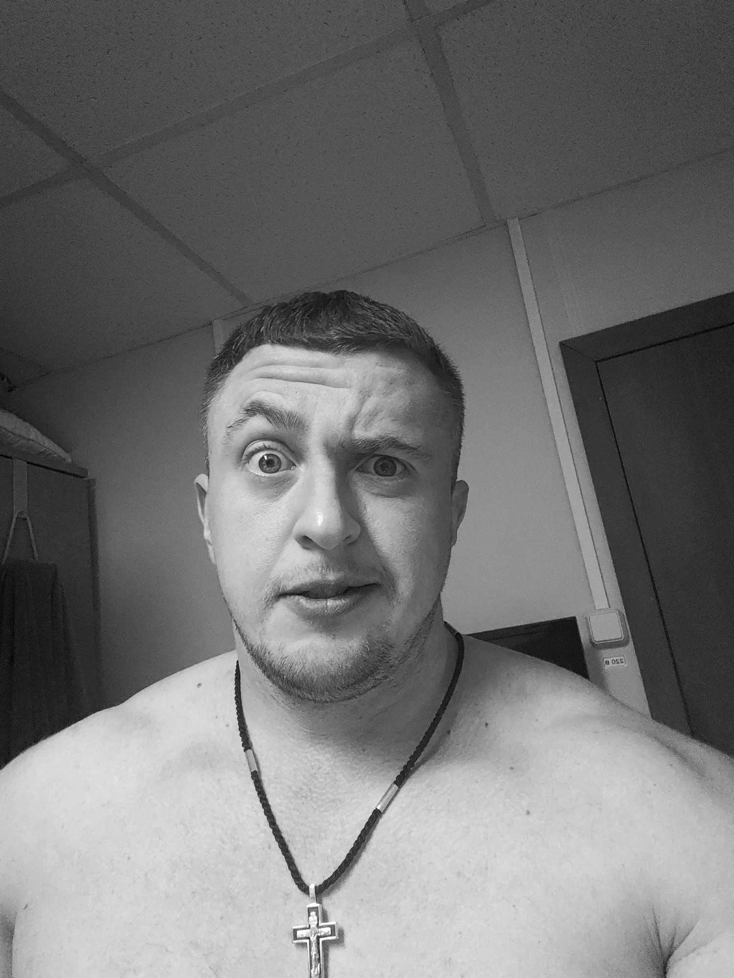 A close-up, grayscale image depicts a shirtless man with a concerned expression, wearing a necklace with a cross pendant. He has short hair and a stubbly beard. The background includes a ceiling with rectangular panels, a wall, a door, and what appears to be a light switch or outlet. The location is unspecified.