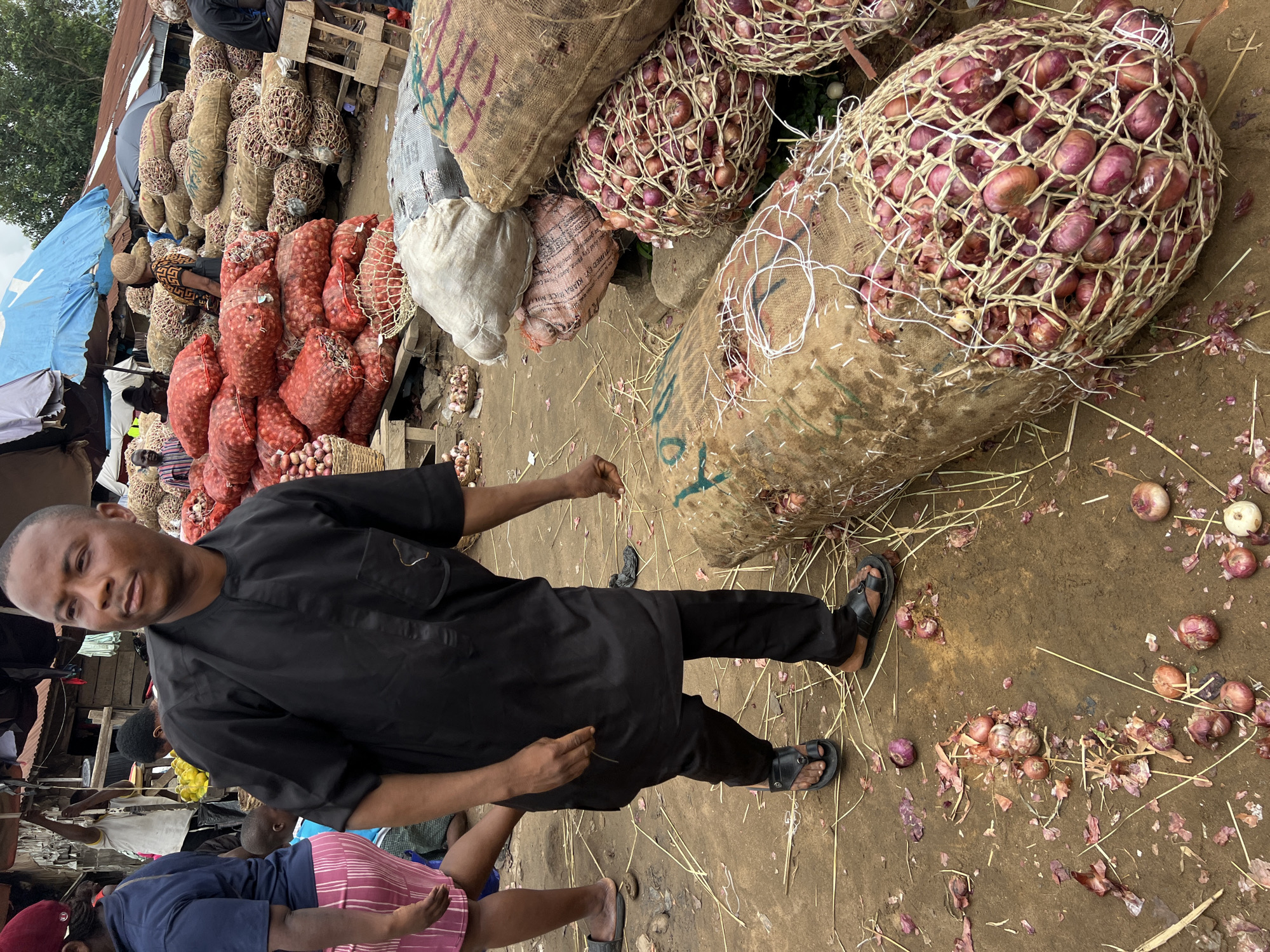 The image depicts an outdoor market scene in Asaba, Nigeria, likely a wholesale produce market. In the foreground, a man wearing a black traditional African outfit with a mandarin collar and black trousers walks across a dusty ground covered with straw and scattered onion peels. He is wearing black sandals. To his left, the legs of a person in a blue shirt and a pink and white striped skirt are visible.
Dominating the background are large piles of onions, predominantly red onions, packaged in burlap sacks and netting. Some sacks are larger, possibly containing other produce like yams, as indicated by a sack with a visible rough texture. Various market stalls with tin roofs and tarpaulin coverings are visible in the distance. The overall impression is of a bustling commercial environment focused on the sale of agricultural goods.
