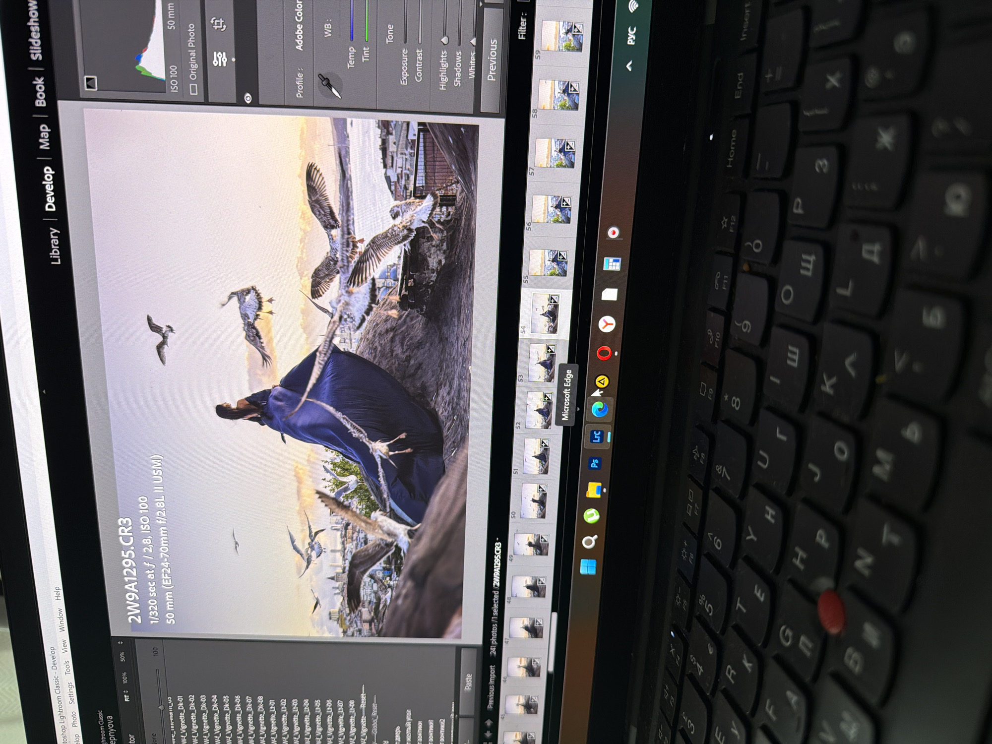 The image shows a laptop screen displaying Adobe Lightroom Classic software. The main subject on the screen is a photograph of a woman in a blue dress standing on a rooftop with seagulls flying around her. The photograph appears to be taken at sunset with the city skyline in the background. The software interface indicates camera settings such as "1/320 sec at f/2.8, ISO 100" and a lens focal length of "50 mm (EF24-70mm f/2.8L II USM)". There is a filmstrip of thumbnail images at the bottom of the screen, and a toolbar at the very bottom shows icons for various applications, including "Microsoft Edge". The laptop keyboard is visible in the foreground. There is no visual information in the image that places the scene in Kadıköy, Turkey.