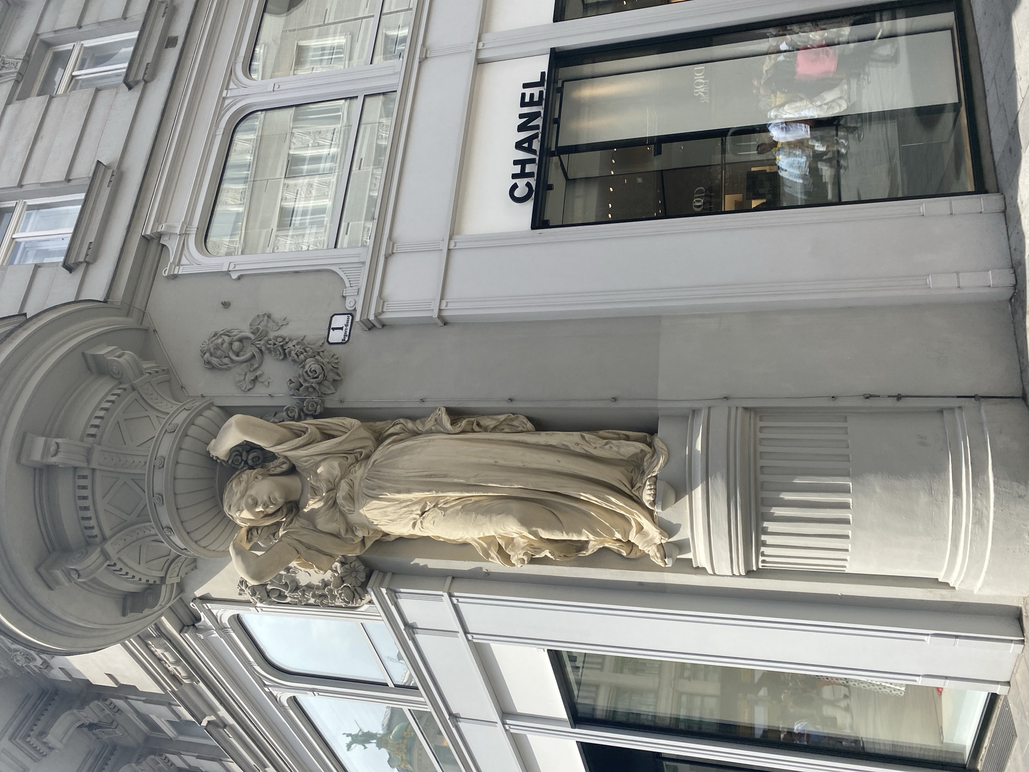 The image shows the exterior of a building in Vienna, Austria, featuring classical architectural elements. A stone statue of a draped female figure holding a floral garland adorns the corner of the building. Above the statue, ornate architectural detailing can be seen, including carvings of flowers and a lion's head. To the right of the statue, a street sign indicates "1, Bognergasse." Below the architectural embellishments, a large storefront displays the "CHANEL" logo in prominent black lettering. The window of the Chanel store reflects the surrounding buildings and passersby. The building appears to be located in a commercial or shopping district, given the presence of the luxury brand store. The overall impression is one of elegant and historic urban architecture.