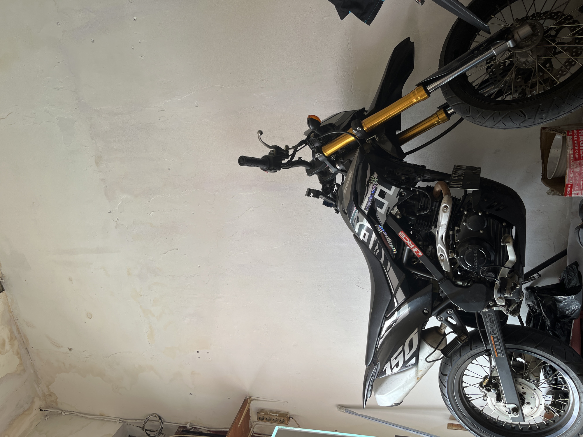 A black Kawasaki dirt bike is parked indoors against a white wall in what appears to be a garage or workshop. The wall shows signs of water damage and discoloration, particularly in the upper left corner. A wooden shelf is mounted on the wall to the left, with electrical wiring and an outlet visible. The motorcycle has gold-colored front suspension forks and various decals on its body, including the brand "Kawasaki" and the word "talapow". A license plate that reads "AD 5580 KI" is attached to the rear. The floor is cluttered with some black trash bags and a cardboard box containing a white bowl. The lighting suggests an indoor setting with some natural light source from the left.