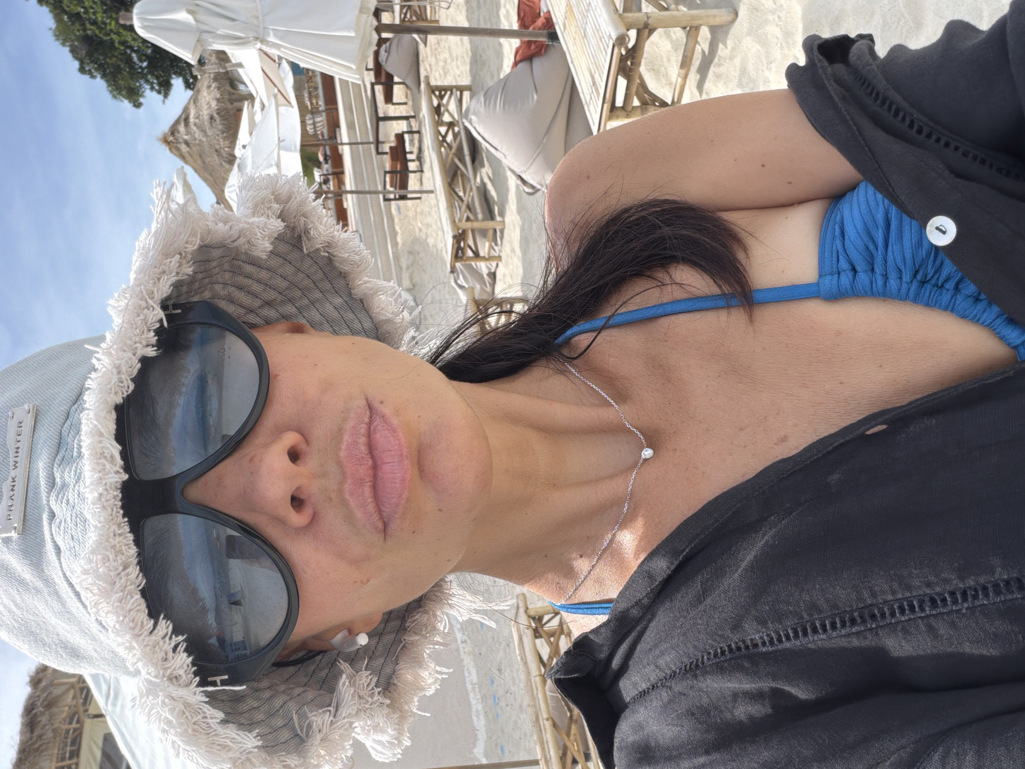 A woman is taking a selfie on a beach in Baan Chaweng, Thailand. She is wearing a light-colored bucket hat with a fringed brim, large dark sunglasses, and a blue bikini top. A black open shirt is draped over her shoulders. She has a delicate silver necklace with a small diamond pendant. The background shows a sandy beach with lounge chairs, umbrellas, and thatched-roof structures. The sky is clear and blue.