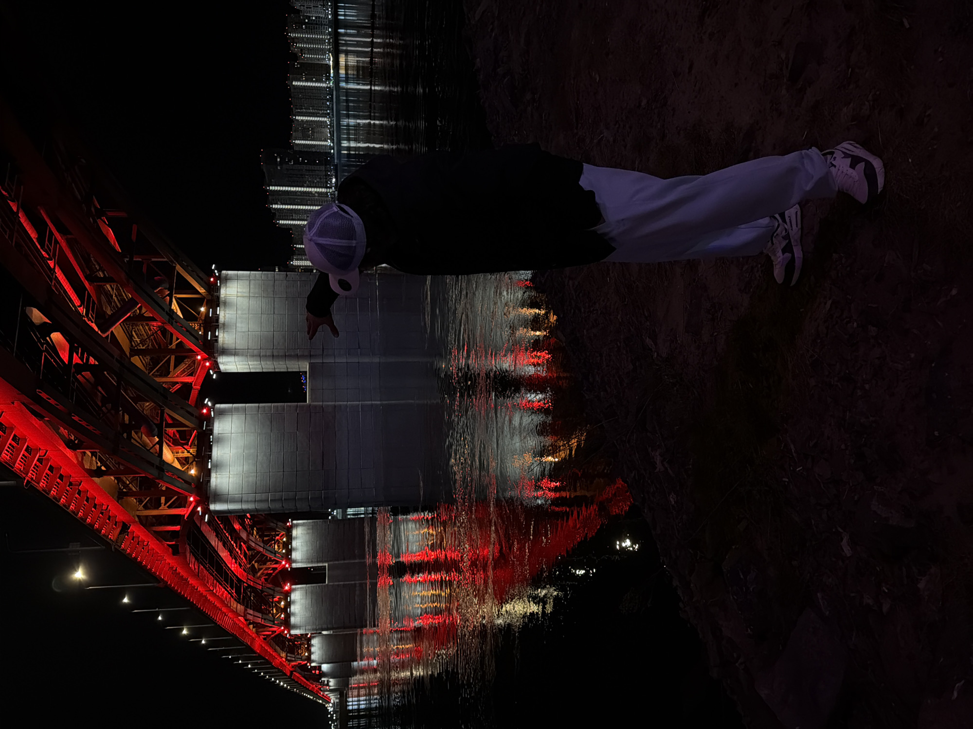 The image depicts a person standing near the Yenisey River in Krasnoyarsk, Russia, at night. The person is wearing a dark jacket, light-colored pants, and a light-colored hat, and they are facing the water. The Krasnoyarsk Railway Bridge is visible in the background, illuminated with red lights. The water reflects the lights of the bridge and the surrounding city. The ground near the person appears to be dirt or soil.