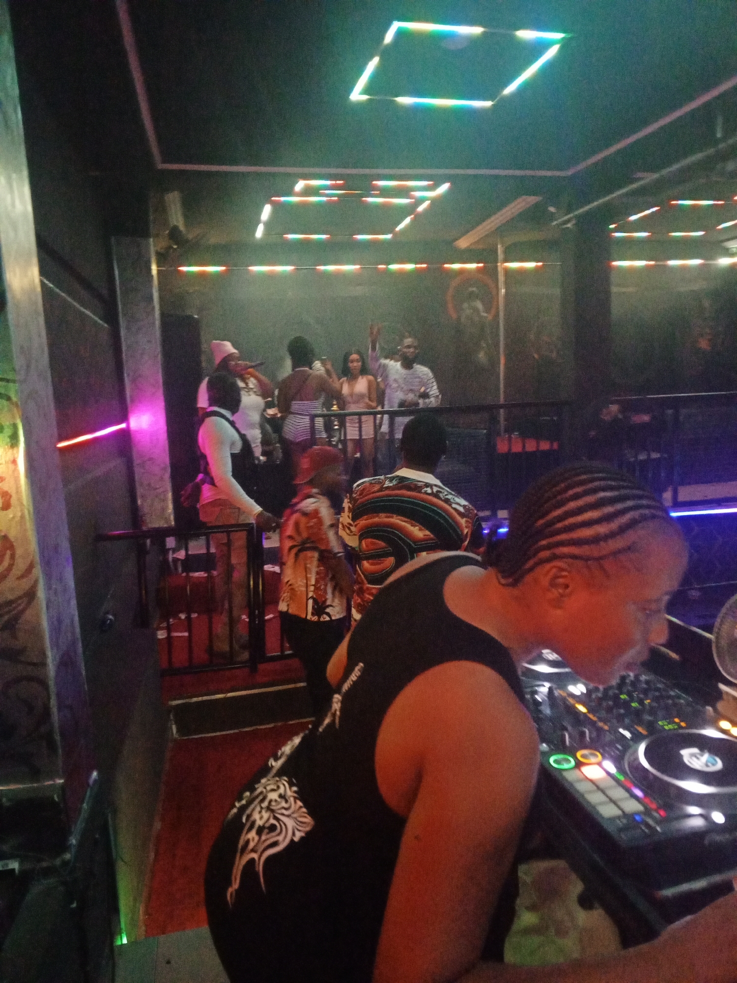 The image shows a dimly lit interior scene, possibly a nightclub or bar, with multiple people present. In the foreground, a person with braided hair is positioned at a DJ setup with various buttons and knobs. The person is wearing a black tank top. In the background, there's a raised platform with several people standing and socializing. At least one person is holding a microphone. The lighting is colorful, with light strips and rectangular fixtures on the ceiling illuminating the room. There are metallic pillars on the left side, and an artwork or mural is faintly visible on the wall behind the platform. The overall atmosphere suggests a lively and social gathering.