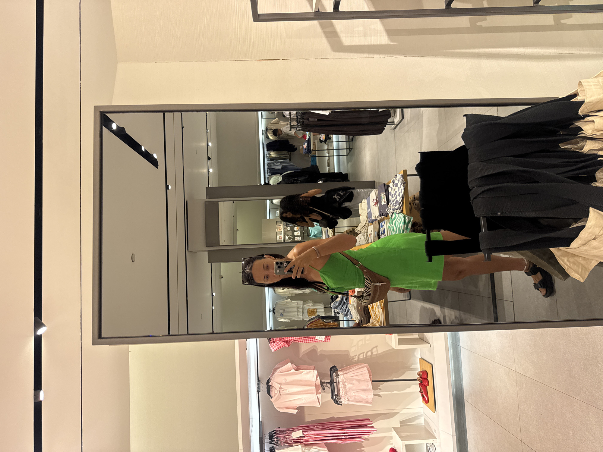 A woman in a bright green slip dress and black sandals is taking a mirror selfie in a clothing store. She is wearing sunglasses on her head and a small brown striped crossbody bag. In the reflection of the mirror, the store's interior is visible, including racks of clothing, display shelves, and other shoppers. To the left of the woman, there are racks of pink clothing. In the background, other shoppers can be seen browsing the merchandise. The store appears to be well-lit and organized. The scene is likely in Kuta, Indonesia, given the context of the prompt.