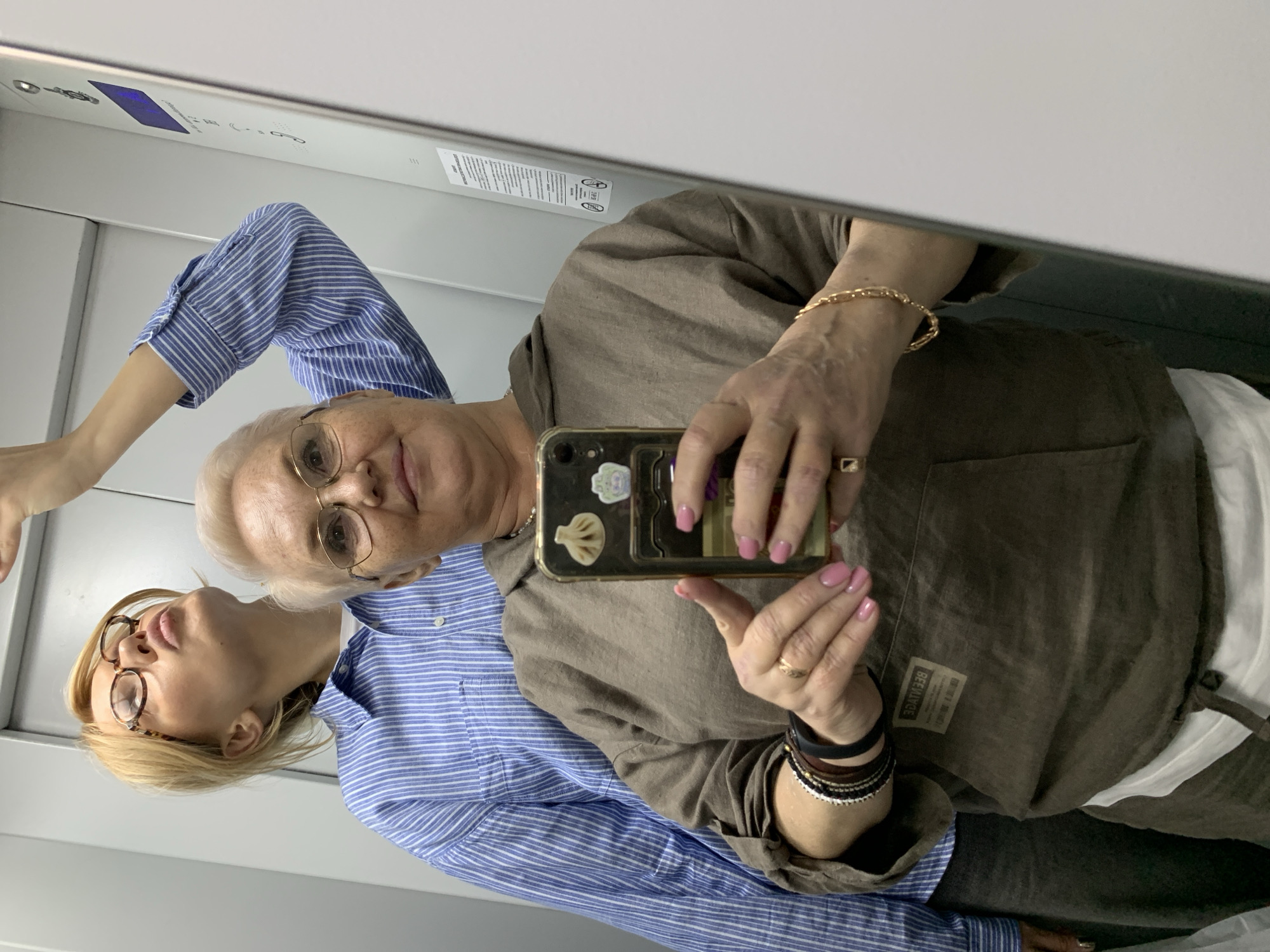 Two women are taking a selfie in a mirrored space, likely an elevator or small room. The older woman is holding a phone, its case adorned with stickers, and has pink nail polish. She wears a brown hooded top and has a gold bracelet and ring. The younger woman is wearing a blue striped shirt. Their faces are visible in the reflection. The background is a plain, light-colored wall. The location is Chaikovsky, Russia.