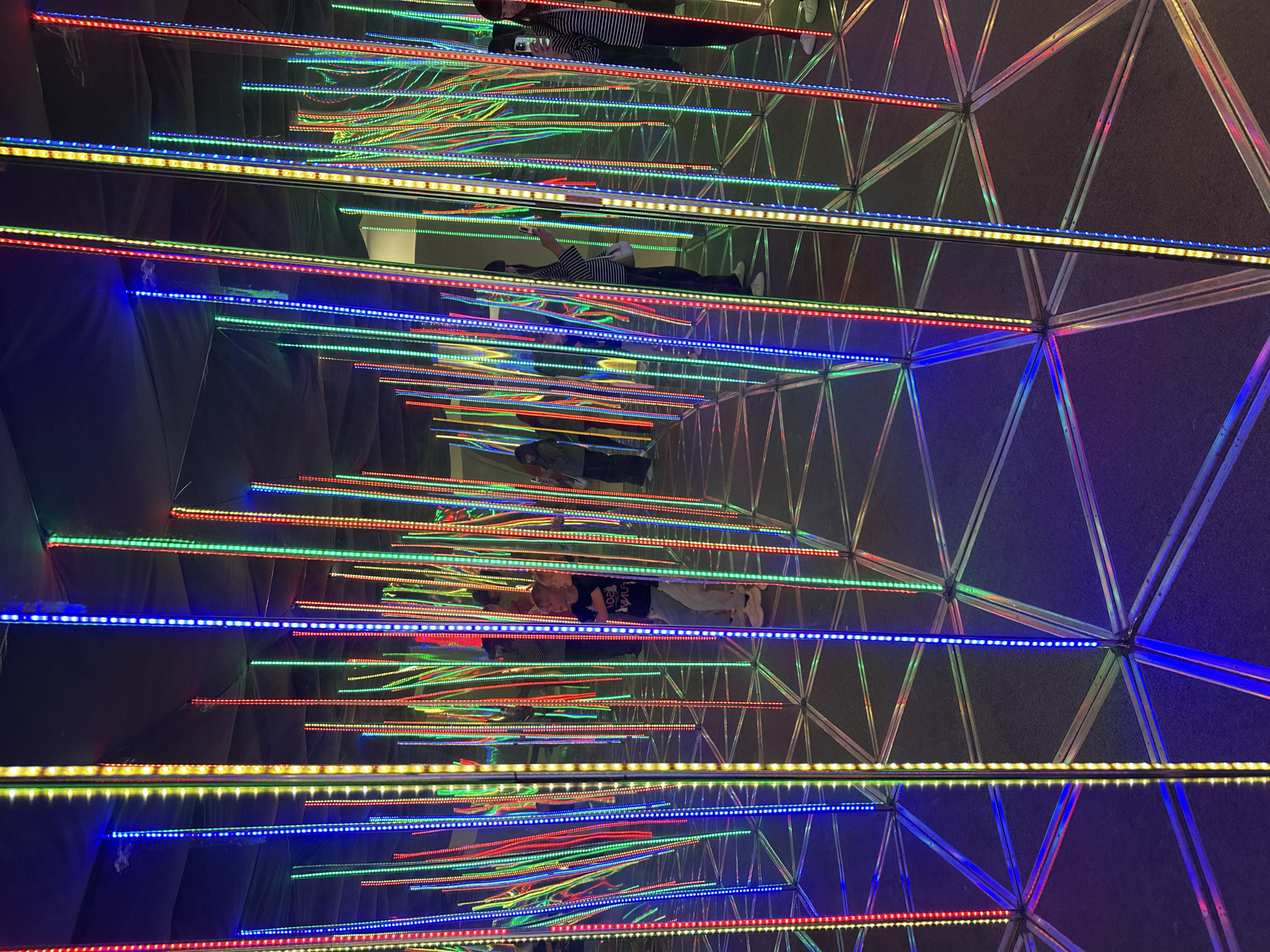 The image depicts an indoor art installation featuring an infinity mirror room. Numerous vertical LED light strips in red, green, blue, and yellow are arranged densely, creating a kaleidoscopic effect through reflections. The floor is a geometric pattern, likely formed by the reflections of the lights and mirrors. Several individuals, including children and adults, are visible within the installation, appearing to interact with or observe the lights. One child is looking towards the right, while another is partially obscured. Adults are seen taking photos and moving through the space. The environment is dimly lit, with the LED lights being the primary source of illumination. The overall impression is one of an immersive and visually stimulating experience. The location is identified as the "IZGM" (Izhevsk State Museum of Fine Arts) in Izhevsk, Russia, suggesting this is a contemporary exhibition space.