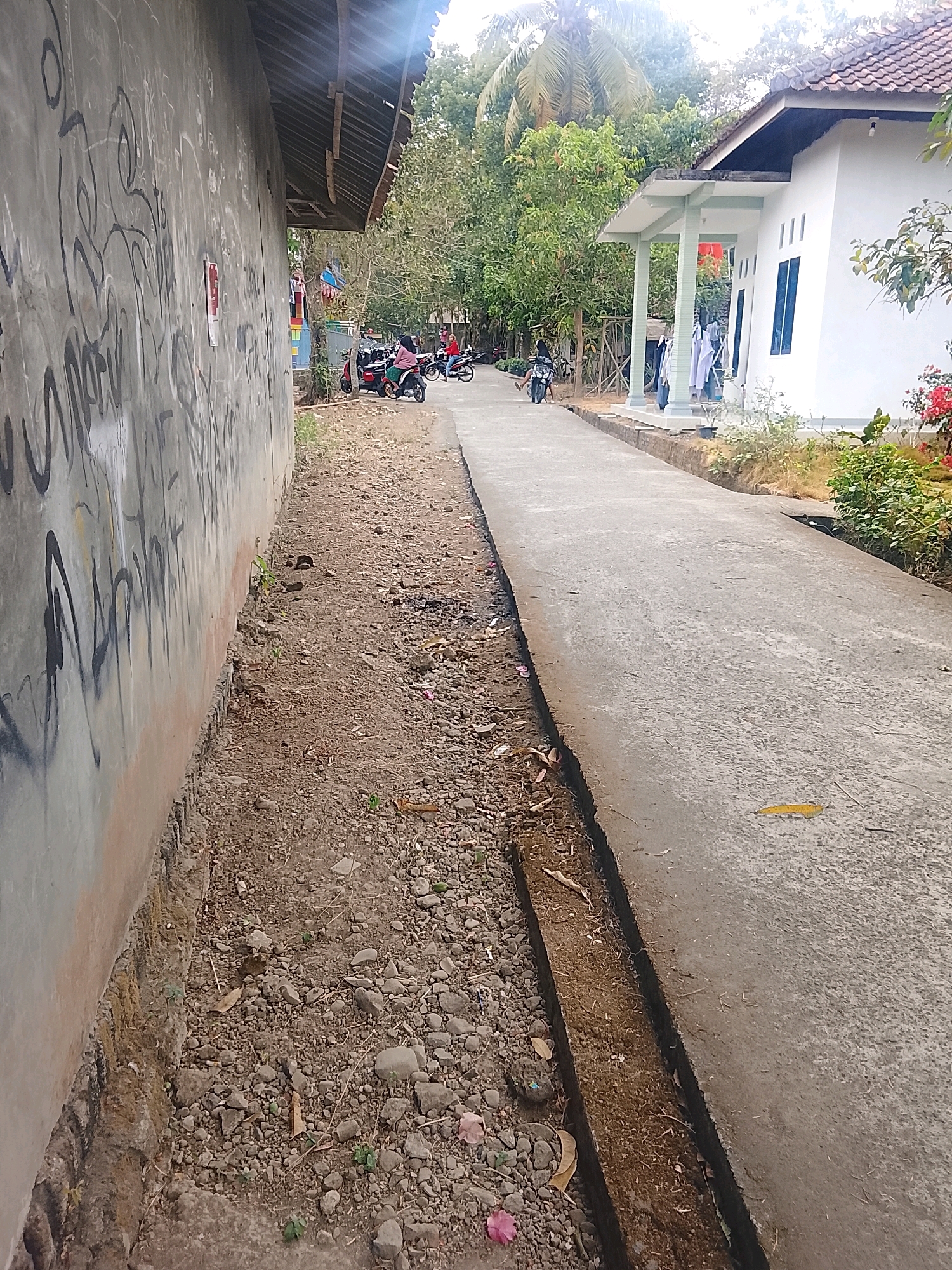 The image depicts a street scene in Kertahayu, Indonesia. On the left, a textured concrete wall with graffiti runs along the side of the frame. Adjacent to the wall is a patch of rough, gravelly ground with scattered dry leaves and small green plants. A paved concrete road occupies the right side of the image, running diagonally into the distance. The road has a distinct trench or ditch running along its edge, filled with dirt and debris.
In the background, several people are visible on motorcycles. Two motorcycles are stopped near the graffiti wall, with individuals standing next to them. Further down the road, more motorcycles are seen in motion, suggesting a common mode of transportation. To the right of the road, a white house with a tiled roof and a covered porch supported by pillars stands. Clothes are hung out to dry on a line near the porch. Lush green trees and foliage surround the house and line the street, indicating a tropical or subtropical environment. The sky is bright with sunlight, casting shadows and creating a slightly overexposed effect in the upper portion of the image. The overall impression is a glimpse of everyday life on a residential street in a rural or semi-rural Indonesian setting.