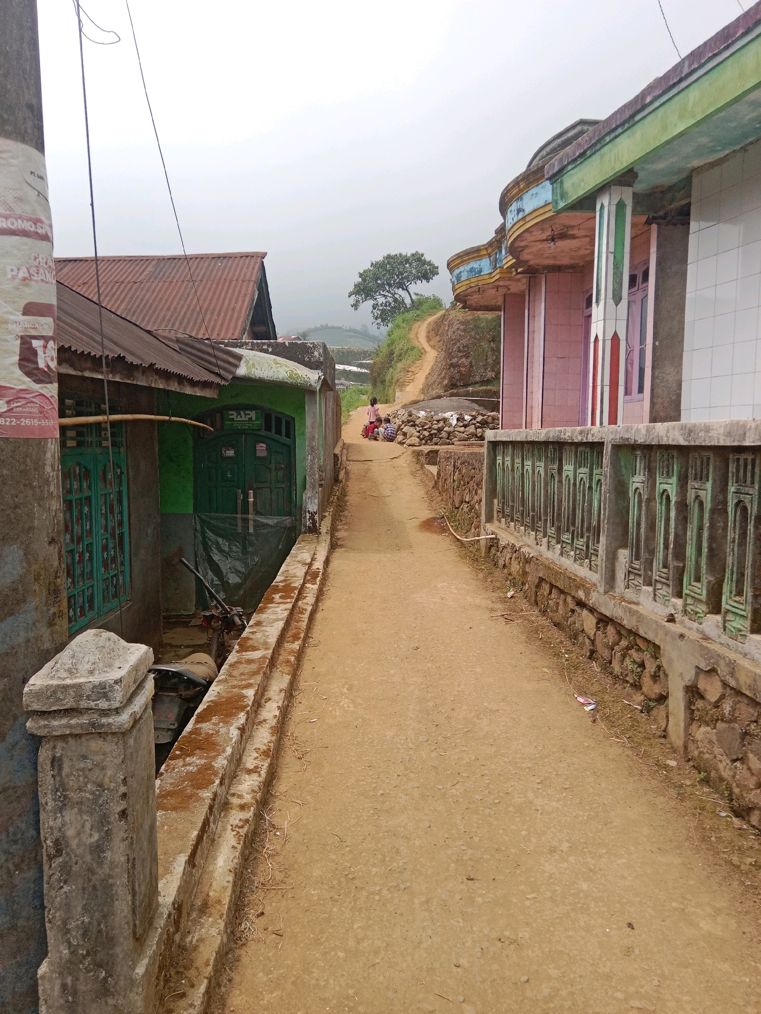 The image depicts a dirt path in a rural area, possibly a village, with buildings on either side. The path slopes upwards and curves to the left, leading towards a single tree in the distance. The sky is overcast. On the left, a green building with a corrugated metal roof is visible, with a motorcycle parked near its entrance. A weathered concrete pillar stands in the foreground of this building. On the right, a pink building with decorative columns and a green and blue trim is seen, bordered by a decorative concrete railing. Two young girls are seated on the path in the middle distance, facing away from the camera. The environment appears to be a mountainous or hilly region, with terraced fields visible beyond the path. The overall impression is one of a humble, possibly remote, community. The location is identified as Batur, Indonesia.