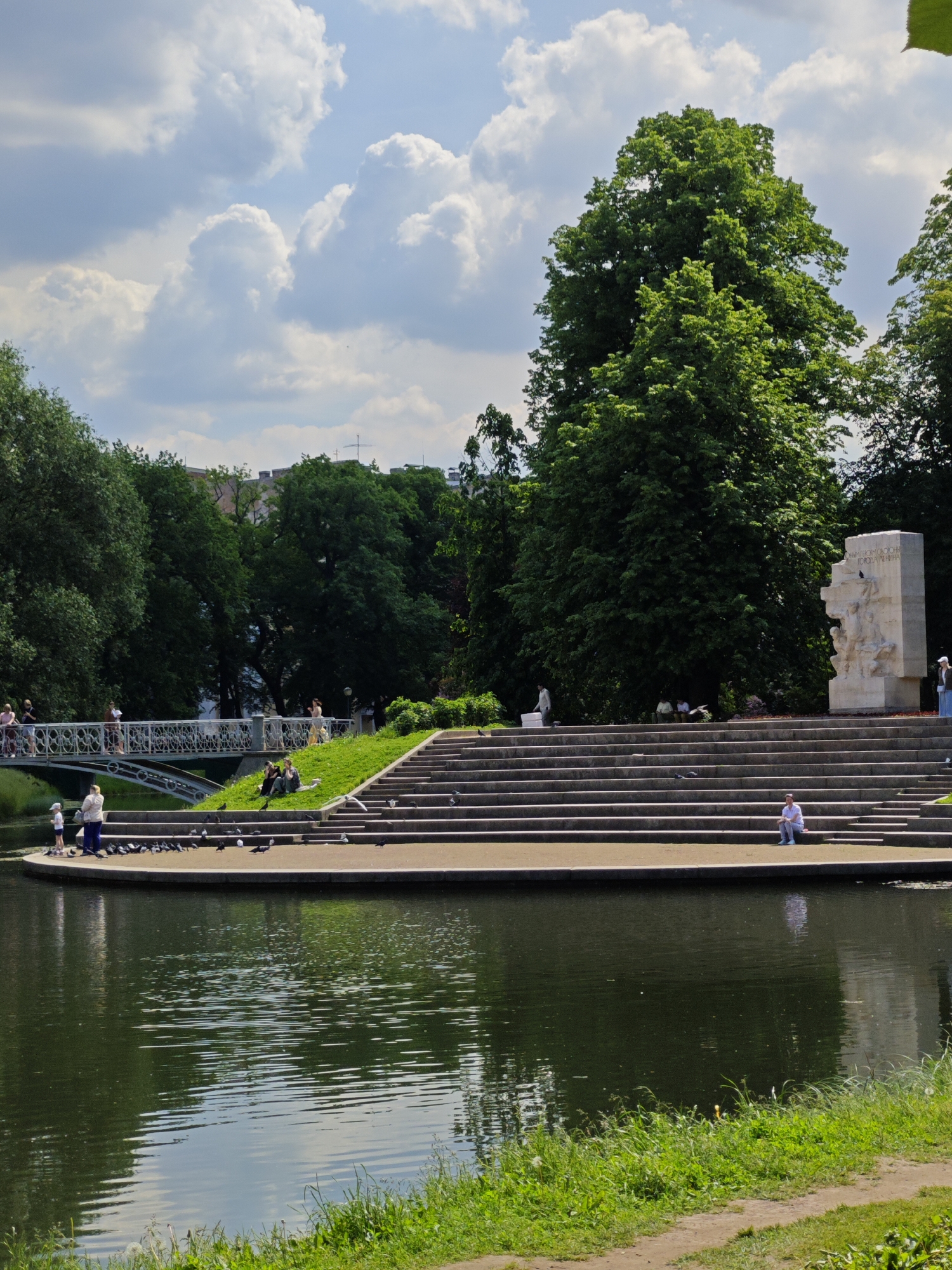 The image depicts a sunny day in a park with a body of water in the foreground. On the far bank of the water, there are stone steps leading up to a grassy area. Several people are visible, some sitting on the steps or grass, others walking or standing. To the left, a white pedestrian bridge spans the water, with more people crossing it. Lush green trees line the background, and a monument, possibly a memorial, stands to the right of the steps. The sky is partly cloudy with large, fluffy white clouds. This scene is characteristic of the Tauride Garden (Tavrichesky Sad) in Saint Petersburg, Russia, a popular public park known for its greenery, water features, and historical monuments.