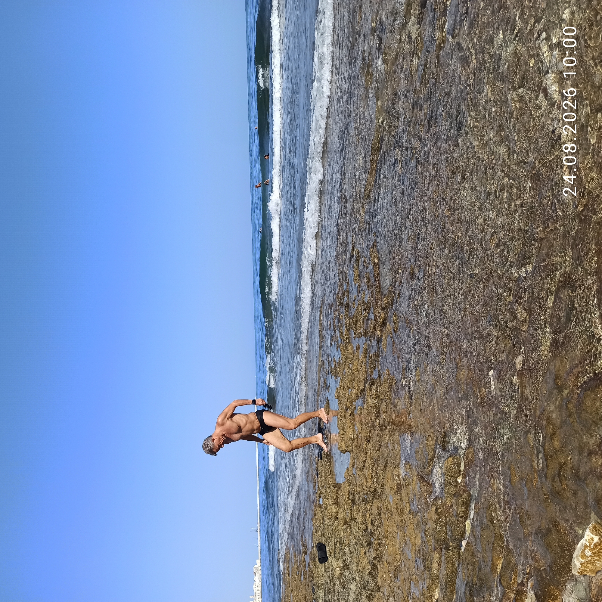 A man in black swim trunks stands on a rocky shore near the water's edge, looking down. He has grey hair and is holding a black object in his right hand. His feet are bare and wet. The water is clear with small waves approaching the shore. In the distance, there are buildings and structures that appear to be part of a city. The sky is clear and blue. The date and time stamp "24.08.2026 10:00" is visible in the lower right corner. This scene is located on the coast of Molfetta, Italy.