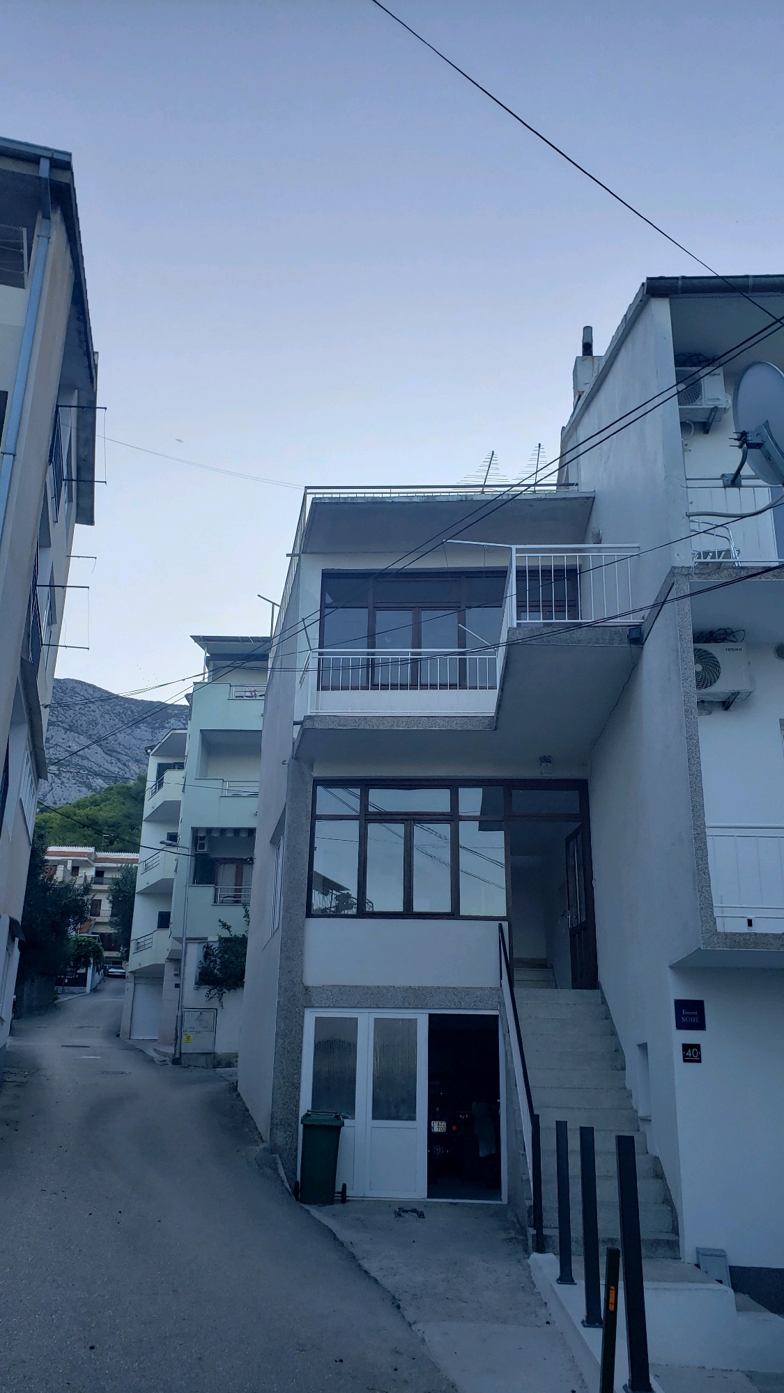 The image shows a street view in Općina Tučepi, Croatia, with several multi-story buildings lining a paved road that slopes upwards. The architecture is consistent with coastal Mediterranean towns, featuring white and light-colored facades, balconies, and visible air conditioning units. On the right side of the image, a white building has a prominent balcony with a white railing on the upper level, overlooking a lower section with large glass windows and an open doorway leading to a staircase. Below this, there is a ground-level garage-like opening with a green trash bin and a white double door. A street sign labeled "Sobe" (meaning rooms, likely indicating lodging) and the number "40" is affixed to the wall of this building. Further up the street, other buildings of similar design are visible, with the backdrop of a lush green hillside and a rocky mountain range under a pale sky. There are no people clearly visible in the frame. The overall impression is a quiet residential or tourist area within a coastal town.