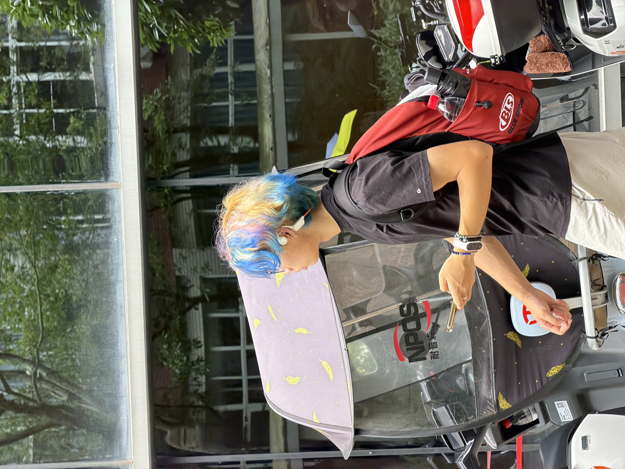 A person with blue and blonde hair is fueling a black and grey electric vehicle in what appears to be the Chongqing High-tech Zone, China. The vehicle has a transparent front windshield with a black sunshade adorned with gold feather patterns. The person is wearing a black t-shirt, light-colored pants, and a red backpack labeled "BLS". The vehicle's fuel filler cap is white with a red cross design. In the background, there is a window reflecting trees and buildings, suggesting an urban environment.