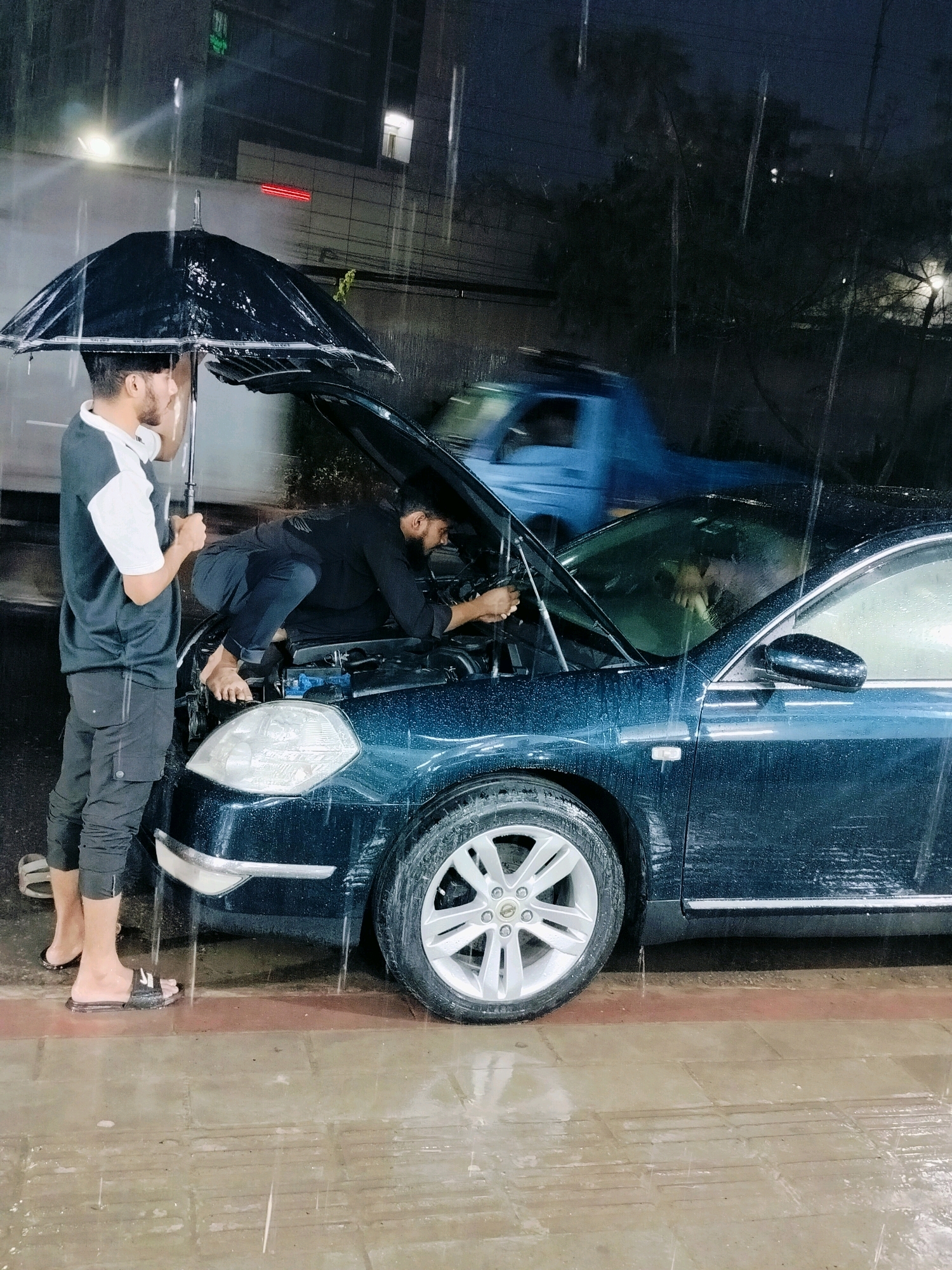 Two men are working on the engine of a dark blue sedan parked on a wet, tiled street at night, with rain falling. One man, wearing a dark shirt and black pants, is crouched over the engine, appearing to be examining or repairing it. The other man, in a black and white polo shirt and black pants, stands beside the car holding a large black umbrella over the engine area, shielding the work from the rain. A blue truck is visible in the background. The scene suggests an improvised repair in adverse weather conditions, possibly in a residential or commercial area of Dhaka, Bangladesh, given the presence of a street and a building in the background.
