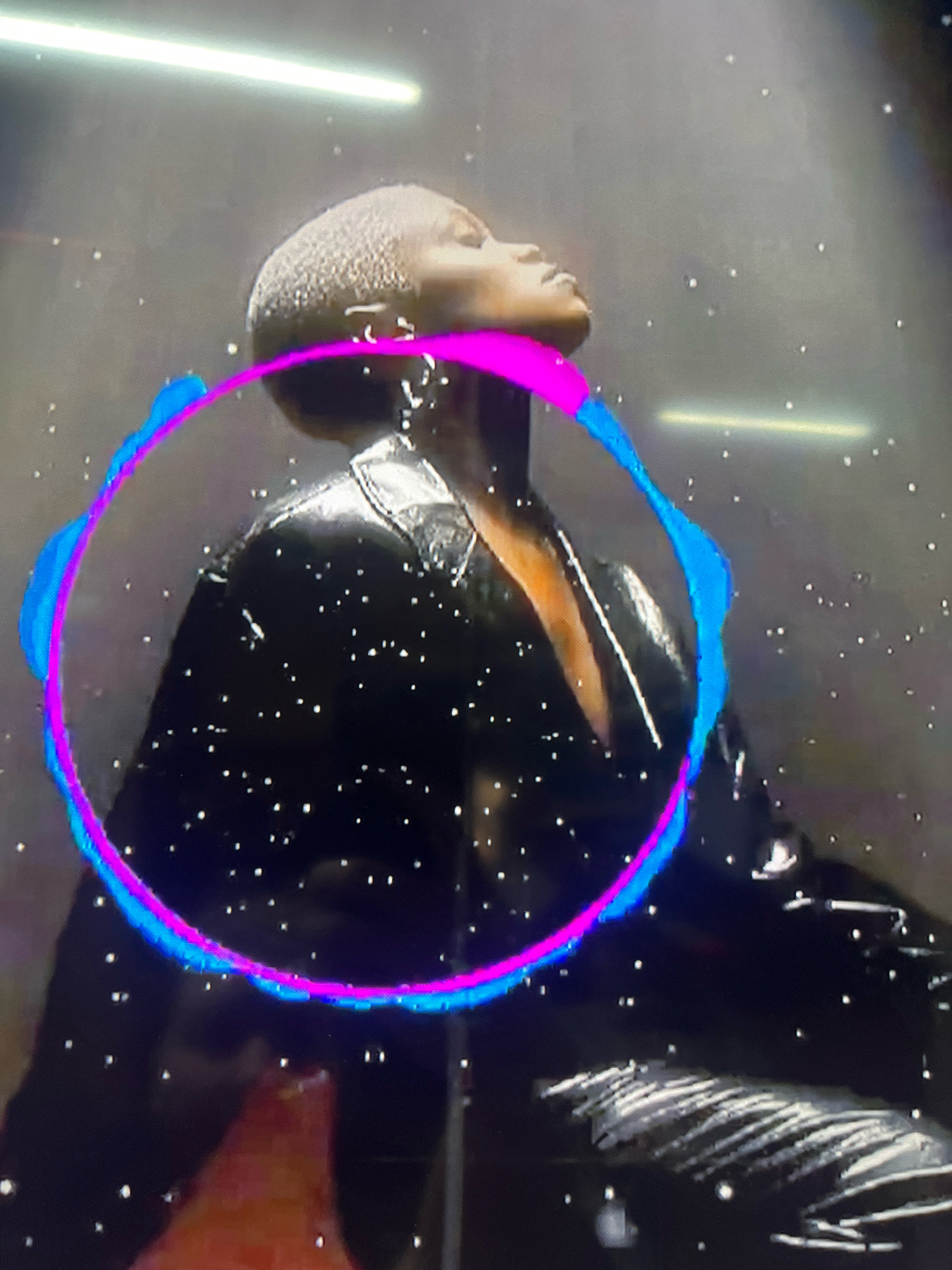 The image is a medium shot of a person with short, buzzed hair, wearing a black, shiny jacket. The person is looking upwards, with their head tilted back. A colorful ring, with magenta and blue hues, encircles the upper body of the person. Small white specks, resembling glitter or light particles, are scattered throughout the image. A bright horizontal light source is visible in the upper background. The background appears to be a dark, possibly smoky or hazy, wall. There are no discernible elements in the image that suggest a specific location in Krasnodar, Russia.