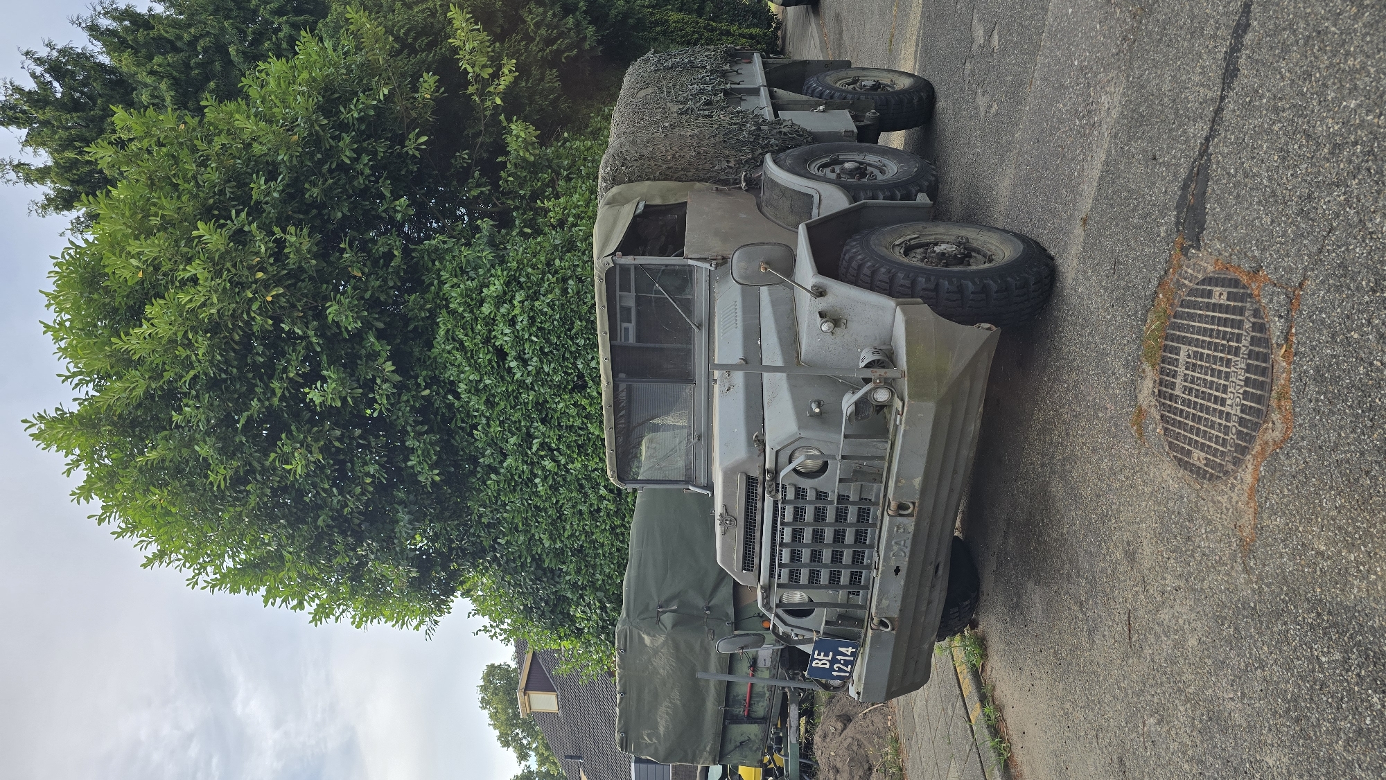 An olive-drab, six-wheeled military truck, possibly a DAF, is parked on an asphalt surface in front of a row of houses and dense green trees. The truck has a canvas covering over its cargo area, which is draped with camouflage netting. The front of the truck features a large grille, a license plate reading "BE 12-14," and a DAF logo. A manhole cover with a grate pattern is visible on the road in the foreground. The sky is overcast with some clouds. The scene appears to be in Zeddam, Netherlands, based on a partial inscription on the manhole cover that reads "VANRINGBOO."
