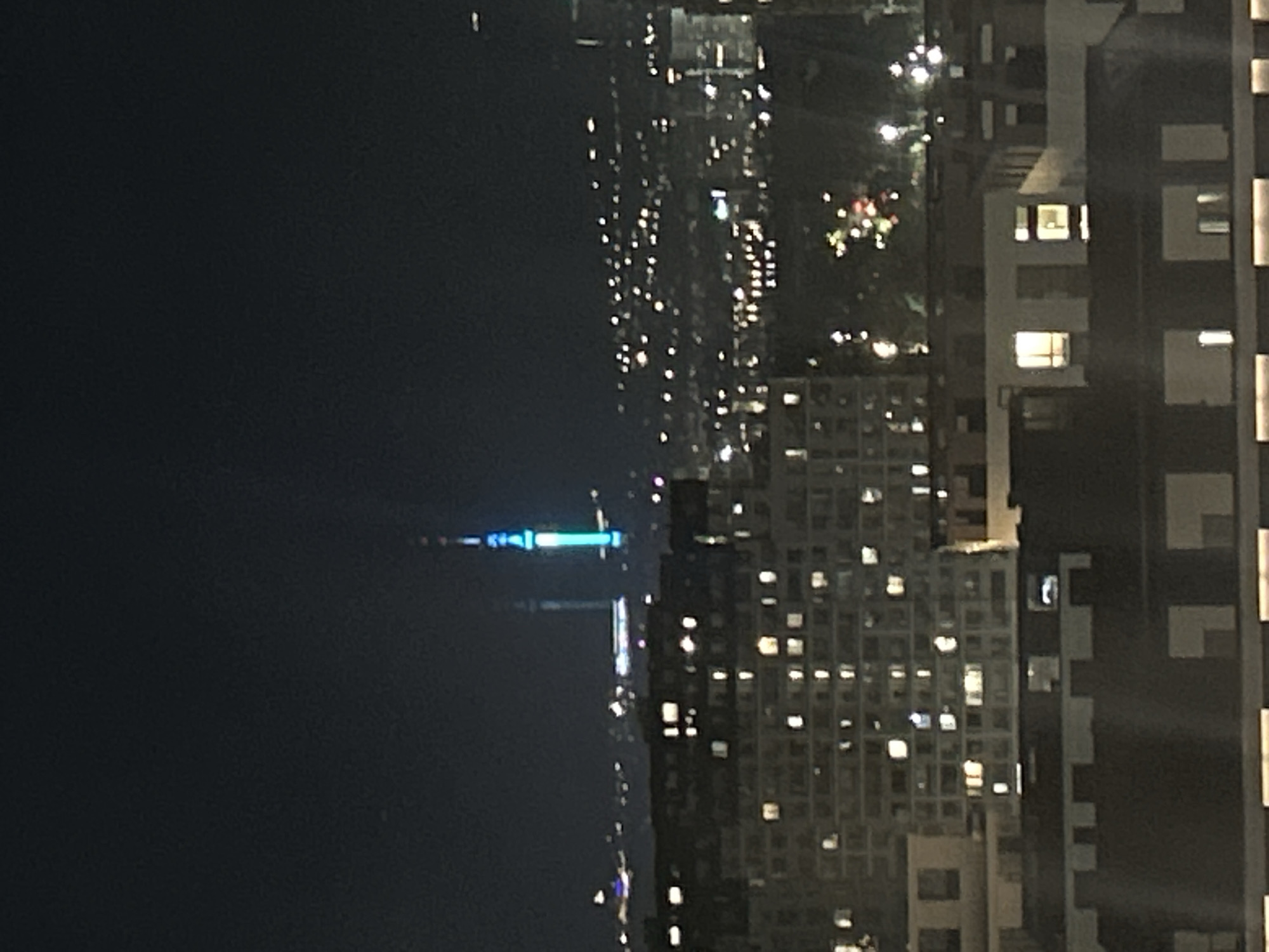 A nighttime aerial view of a cityscape, likely Almaty, Kazakhstan, dominated by illuminated buildings and a prominent, brightly lit tower. The foreground features the dark silhouettes of apartment buildings with numerous windows glowing with warm light, indicating occupancy. Beyond these, the city unfolds with a dense collection of buildings and lights stretching into the distance, suggesting urban sprawl. In the mid-ground and background, a tall, slender tower stands out, illuminated with vibrant blue and white lights, possibly a television or communication tower. Its distinctive illumination makes it a focal point against the dark night sky. The overall scene depicts a lively, well-populated urban environment after dark, with the tower serving as a notable landmark.