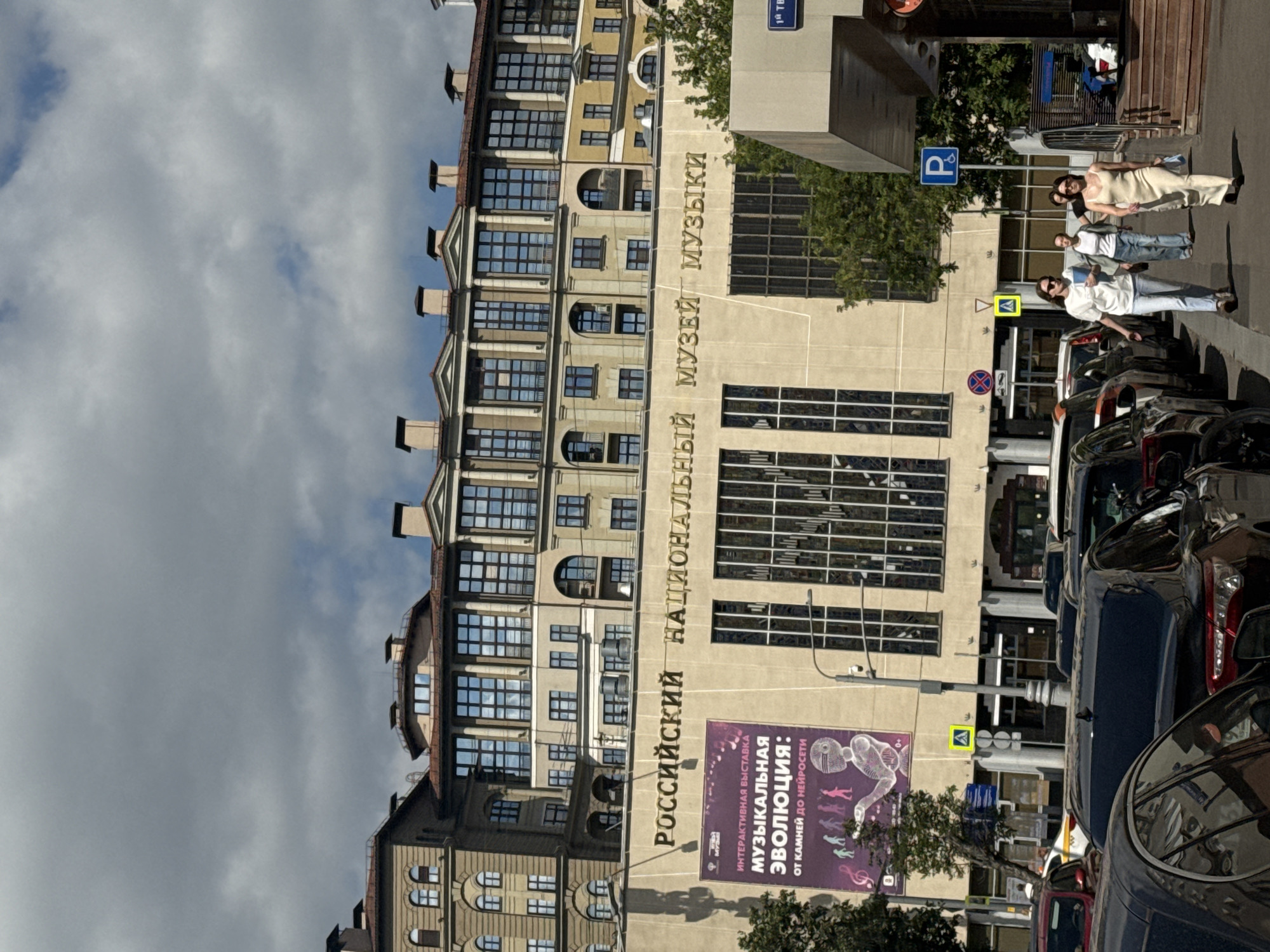 This is a vertical, outdoor photograph of a building with Cyrillic text on its facade and people walking on the sidewalk. The building appears to be a museum, with the text reading "РОССИЙСКИЙ НАЦИОНАЛЬНЫЙ МУЗЕЙ МУЗЫКИ" (Russian National Museum of Music). A poster on the building advertises an interactive exhibition called "МУЗЫКАЛЬНАЯ Эволюция: ОТ КАМНЕЙ ДО НЕЙРОСЕТИ" (Musical Evolution: From Stones to Neural Networks).
Several people are visible on the sidewalk in front of the building, some walking and some standing. One group of three women, dressed in casual summer attire, appears to be posing or gathered together. Another person in a white top and jeans is walking away from the camera.
The setting is urban, with cars parked along the street and a traffic sign visible. The sky is partly cloudy, suggesting it could be daytime with variable weather. The architecture of the building, with its multiple windows and classical-inspired detailing, indicates a potentially older or historically significant structure. The overall impression is a lively street scene outside a cultural institution in Moscow, Russia.