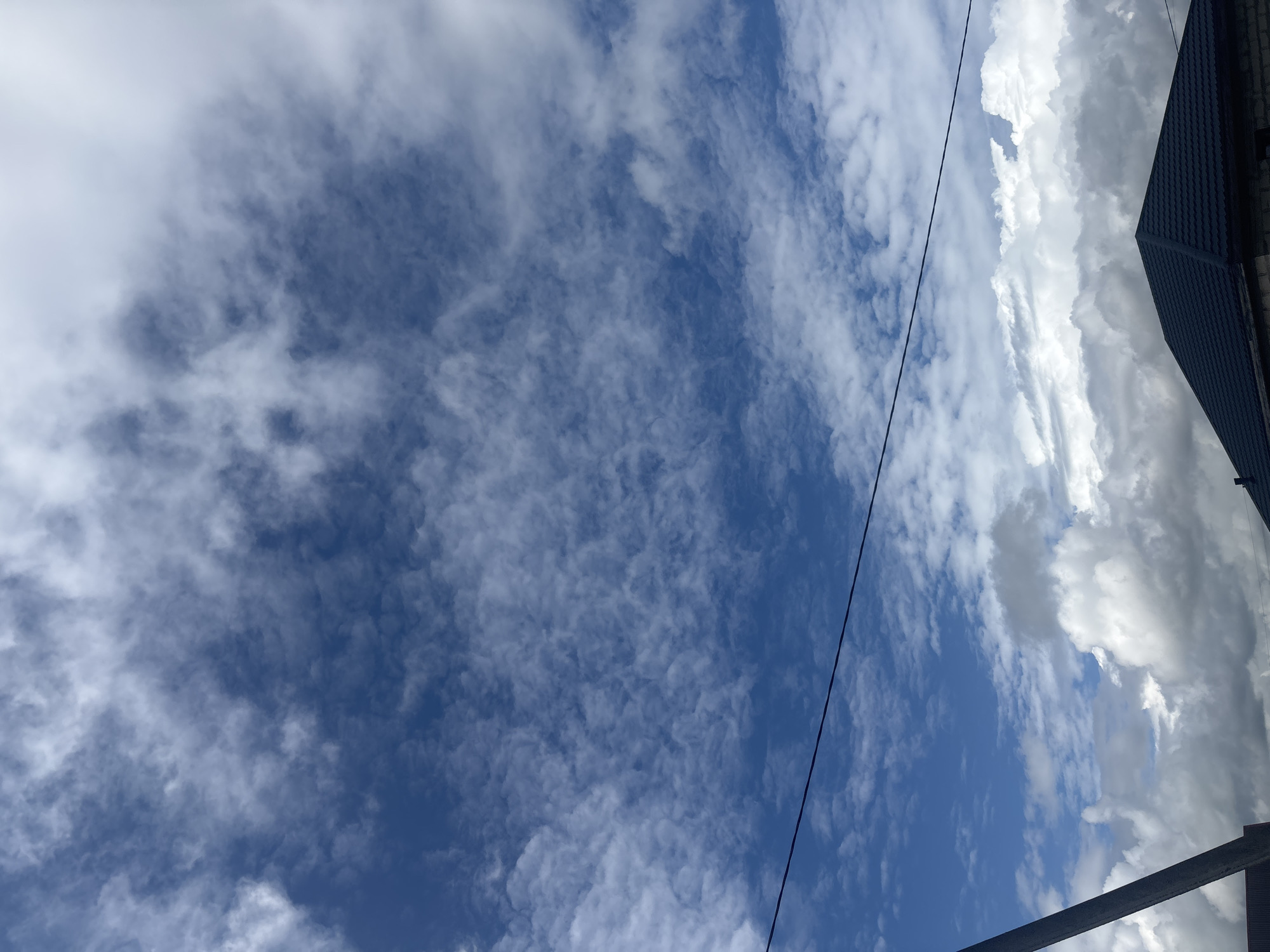 The image depicts a cloudy sky with a dark, angled silhouette of a building's roof in the foreground, possibly a house with a dark grey metal roof. A utility pole is partially visible on the left, and a thin wire stretches horizontally across the upper portion of the frame. The sky is a mixture of deep blue and white clouds, with some appearing wispy and others more dense and fluffy. The overall scene suggests an outdoor environment under a dynamic sky, potentially in a suburban or rural setting. There are no people visible in the image. The location is stated to be in Kapyĺ, Belarus.
