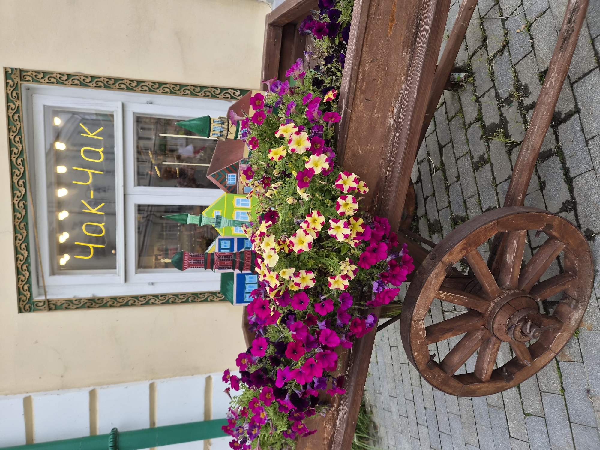 A wooden wagon filled with colorful petunias and miniature decorative buildings sits on a cobblestone street in front of a building with a window. The window has yellow neon lettering that reads "Чак-Чак" (Chak-Chak). This is likely in Kazan, Russia, as Chak-Chak is a traditional Tatar dessert and a common sight in Kazan. The wagon is made of dark wood and has one large spoked wheel visible. The flowers are a mix of vibrant pink, purple, and yellow. The miniature buildings appear to be stylized representations of traditional Russian architecture, with green and red roofs. The building in the background is light yellow with white trim around the window.