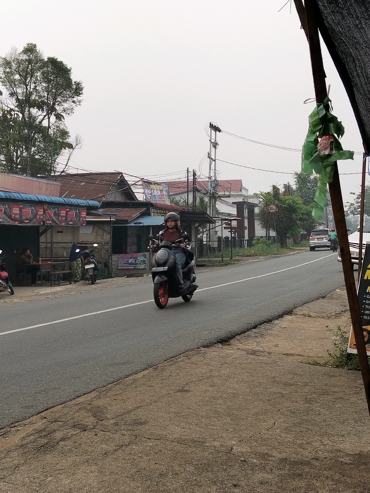 The image captures a street scene in Arang Limbung, Indonesia, on an overcast day. A person wearing a helmet is riding a black scooter down a paved road. The scooter has red accents on its wheels. On the left side of the road, there are several small buildings with corrugated metal roofs, some of which appear to be shops. People are visible inside one of the buildings, seated at tables. Several other parked motorcycles are also present. A white car is seen in the distance on the right side of the road, along with another motorcycle and rider. A speed limit sign indicating "30 km" is visible on a pole. The environment suggests a semi-urban or roadside setting, with vegetation and electrical wires present. A dark, textured fabric is draped over a wooden pole in the foreground on the right, with some green plastic bags tied to it. The road surface shows signs of wear.