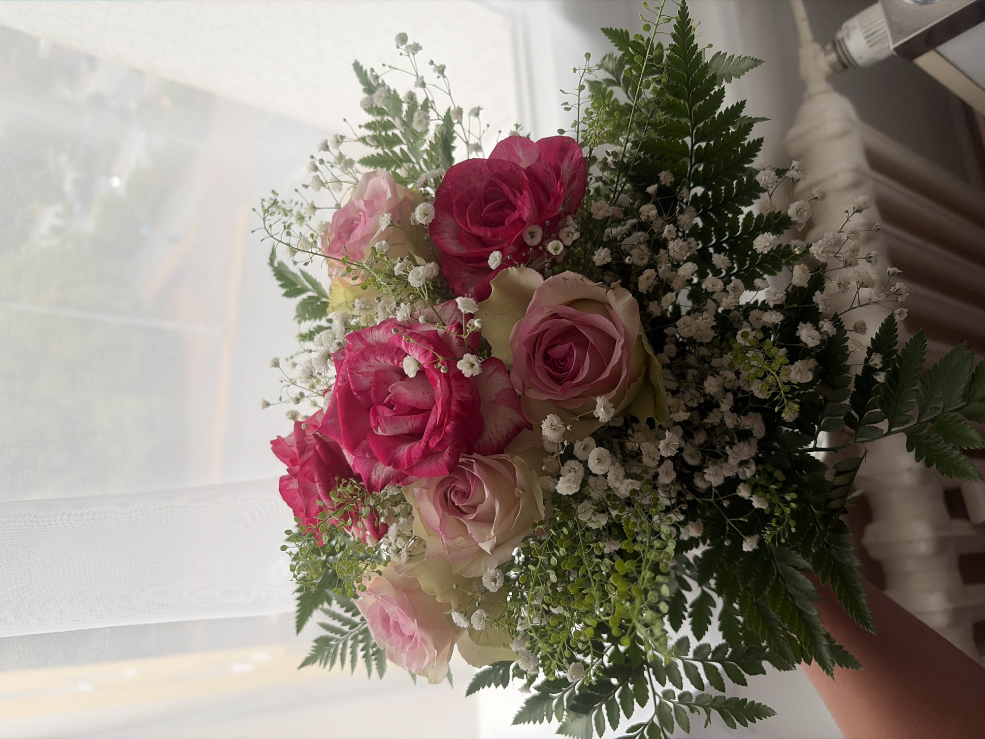 A hand holds a bouquet of roses and baby's breath in front of a sheer white curtain, obscuring a window with a blurry view of trees and buildings, possibly in Duisburg, Germany. The bouquet features a mix of pink and white roses, with some having a marbled or variegated pattern of deep pink. The baby's breath, a filler flower, is scattered throughout the bouquet, creating a delicate, airy effect. Green fern leaves are also interspersed, adding texture and depth. The background is softly lit, suggesting natural light filtering through the curtain. The scene appears to be indoors, with a hint of a radiator visible at the bottom right, further indicating an indoor setting.