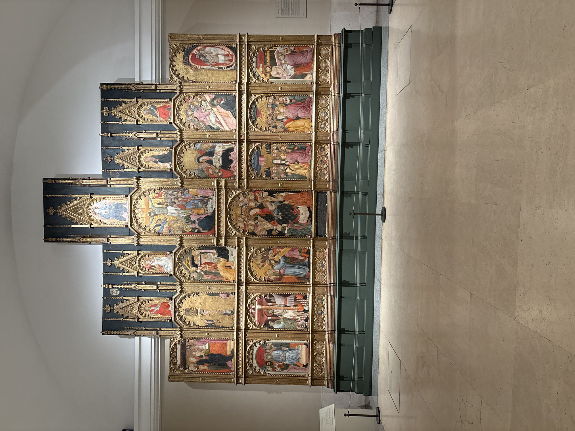 The image shows a large, multi-paneled altarpiece or retablo. The altarpiece features numerous small painted panels, each depicting a religious scene, likely from the life of Christ or a saint. The panels are ornately framed in gold. The altarpiece is positioned on a low green stand and protected by slender black poles. It is located inside a museum with a light-colored stone floor and plain white walls. Based on the visual style and content, it is likely displayed in the Prado Museum in Madrid, Spain. No people are visible.