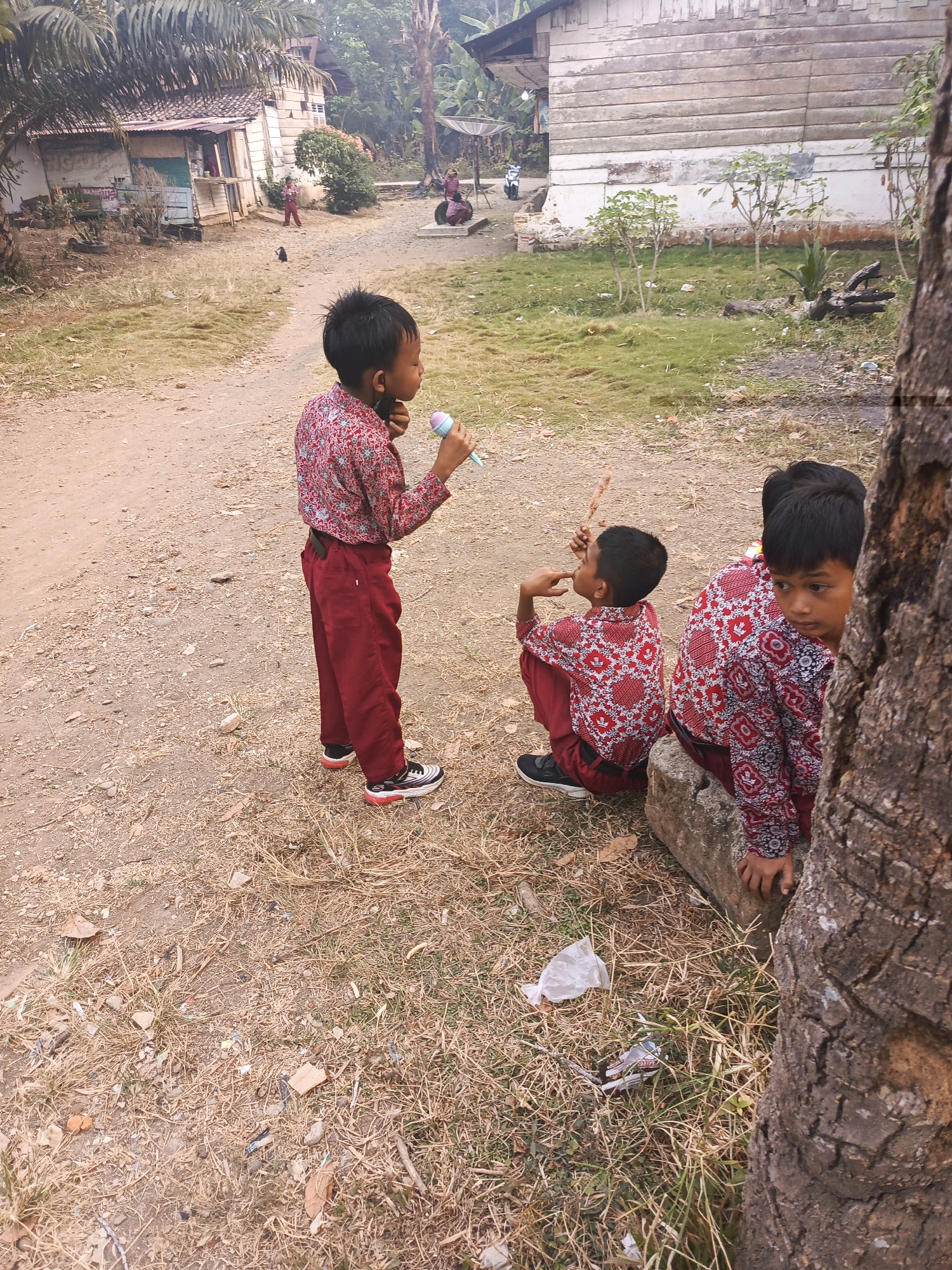 In Megang Sakti II, Indonesia, three young boys in matching red and white patterned shirts and red pants are present. One boy stands facing left, holding a light blue object to his mouth, possibly a toy microphone or a lollipop. Two other boys are crouched down. One is seated on a concrete block and looking towards the right of the frame, while another is seated on the ground to his left, looking upwards and to his left. A large tree trunk is prominently featured on the right side of the image, partially obscuring the view of the boys. The background includes a dirt path, dry grass, and a rustic building with a corrugated metal roof. In the distance, more buildings and vegetation are visible.
