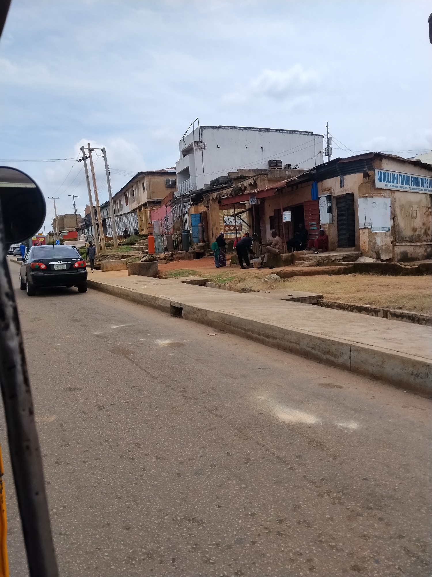 The image depicts a street scene in Jos, Nigeria. A black Toyota Corolla is parked on the right side of the asphalt road, facing away from the camera. To the right of the road, there is a sidewalk with a concrete curb and some dry grass. Behind the sidewalk, a row of buildings lines the street. These buildings appear to be a mix of commercial establishments and residential structures, with some showing signs of wear and tear. One building has a blue sign that reads "ABDULLAHI TAIWO FURNITURE W". Several people are visible, some standing and others seated outside the buildings, suggesting a commercial or community gathering area. Utility poles with electrical wires are present, indicating the presence of public infrastructure. The sky is partly cloudy. The perspective suggests the photo was taken from a moving vehicle.