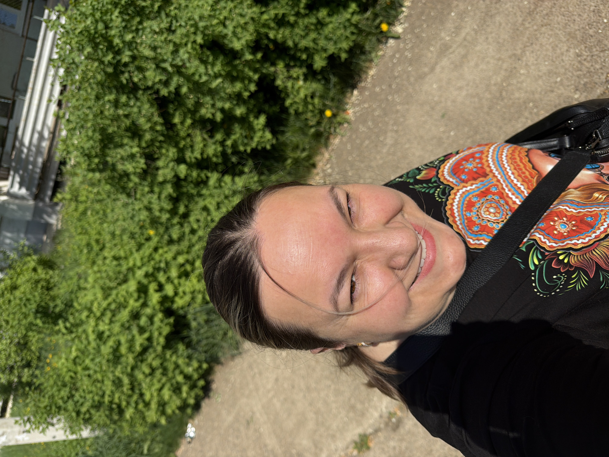A woman is seen smiling in an outdoor photograph. She has light skin, brown hair with blonde highlights, and is wearing a black top with an orange and blue floral pattern. A black bag strap is visible over her shoulder. In the background, there is a green bush and a concrete pavement. The location appears to be in a city park, but the city of Chaikovsky, Russia, cannot be confirmed based solely on the image.