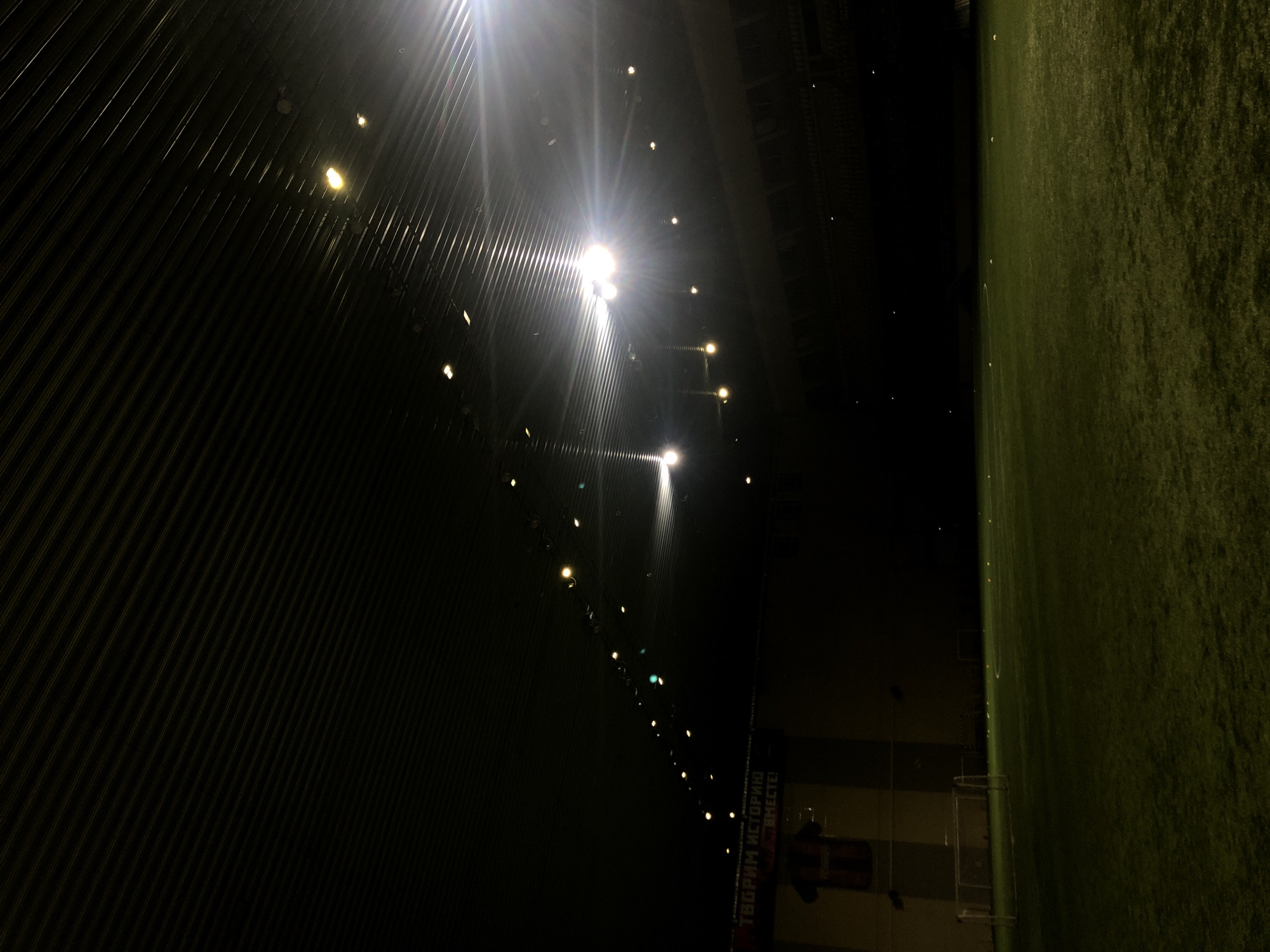 The image shows a view of a football stadium at night. Artificial lights illuminate the field and the surrounding infrastructure. On the left, corrugated metal panels form a wall. On the right is a green field of grass. In the lower portion, a partially obscured banner reads "ТВОРИМ ИСТОРИЮ ВМЕСТЕ!". It is the Yenisey Stadium in Krasnoyarsk, Russia.