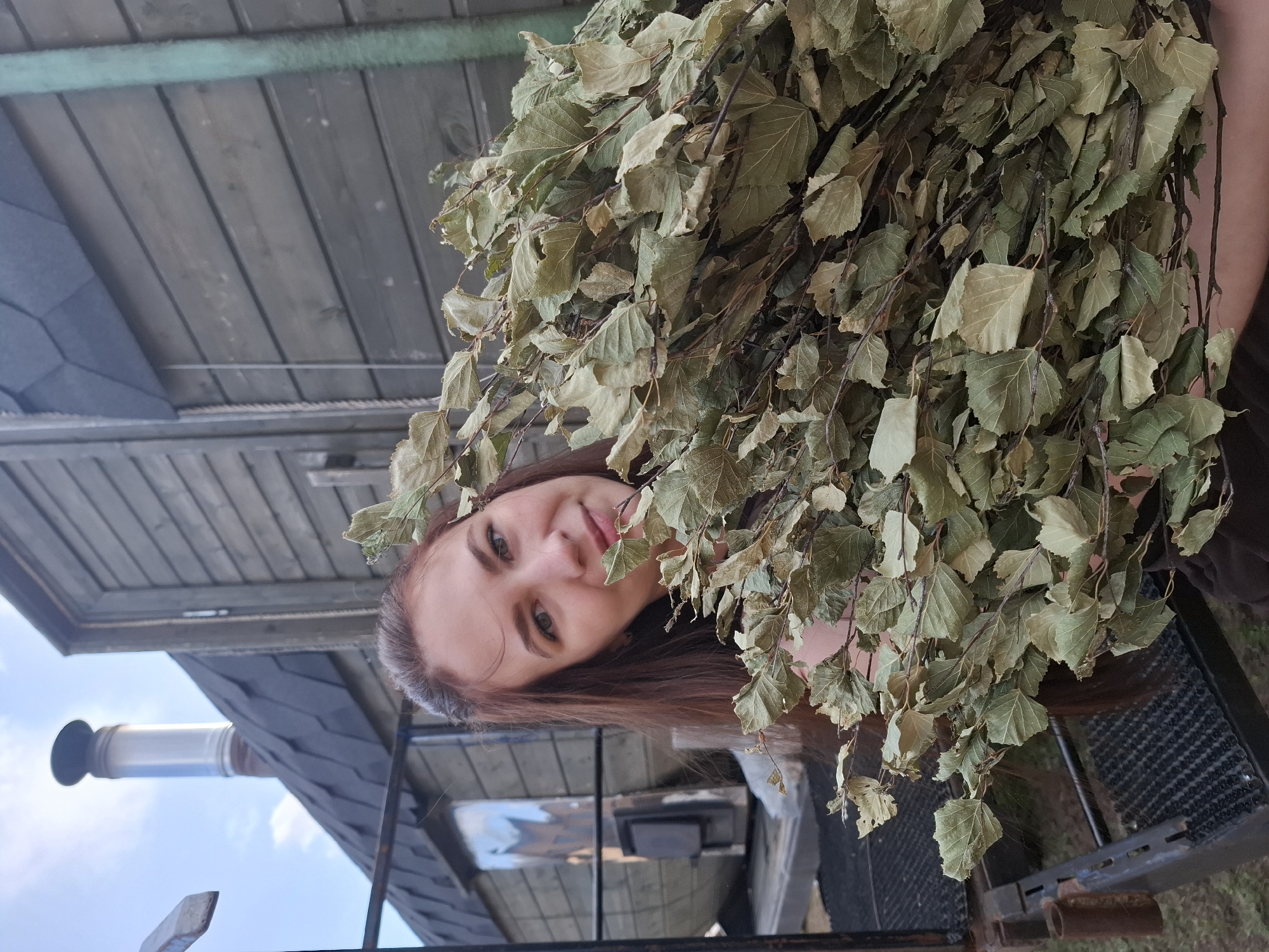 A woman is holding a birch broom in Chaikovsky, Russia. The image is taken from a low angle, looking up at the woman. She has long brown hair and is smiling slightly. The birch broom is large and covers much of the frame. Behind the woman, there is a building with a wooden exterior and a chimney. The sky is visible in the background.