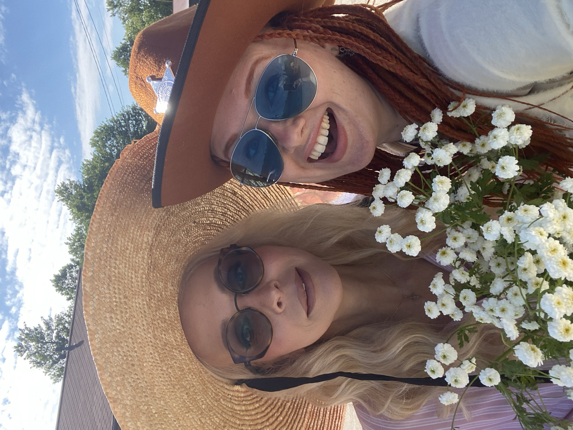 Two women are taking a selfie outdoors, with trees and a building in the background. The woman on the left wears a wide-brimmed straw hat, round sunglasses, and a pink and white striped top. She has long blonde hair. The woman on the right wears a brown cowboy hat with a silver star emblem, aviator sunglasses, and a white fuzzy jacket. She has long, reddish-brown braided hair. Both women are smiling. The woman on the right is holding a bouquet of small white flowers. The image appears to be taken during the daytime, with a blue sky and clouds visible.