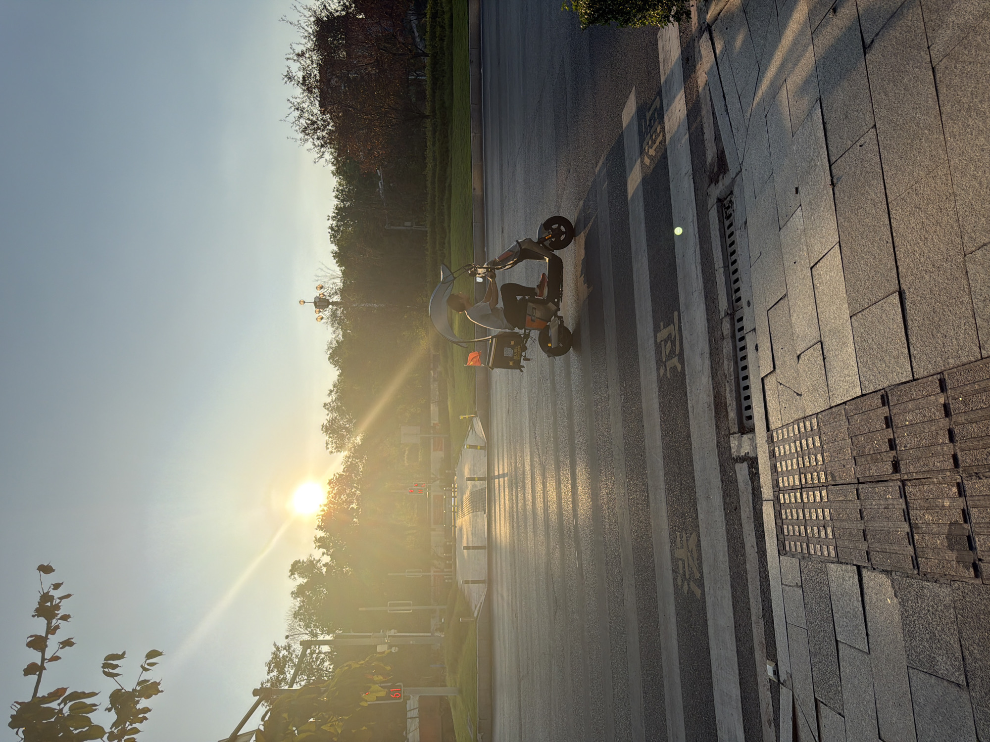A person rides a scooter across a crosswalk in Jiangjin, China, bathed in the golden light of a setting sun. The scooter has a canopy overhead and a black delivery box on the back. The crosswalk is on an asphalt road, with a sidewalk made of stone tiles to the right. In the background, trees and buildings are visible, with a traffic signal and streetlights also present. The sun is low in the sky, casting long shadows and creating lens flare.