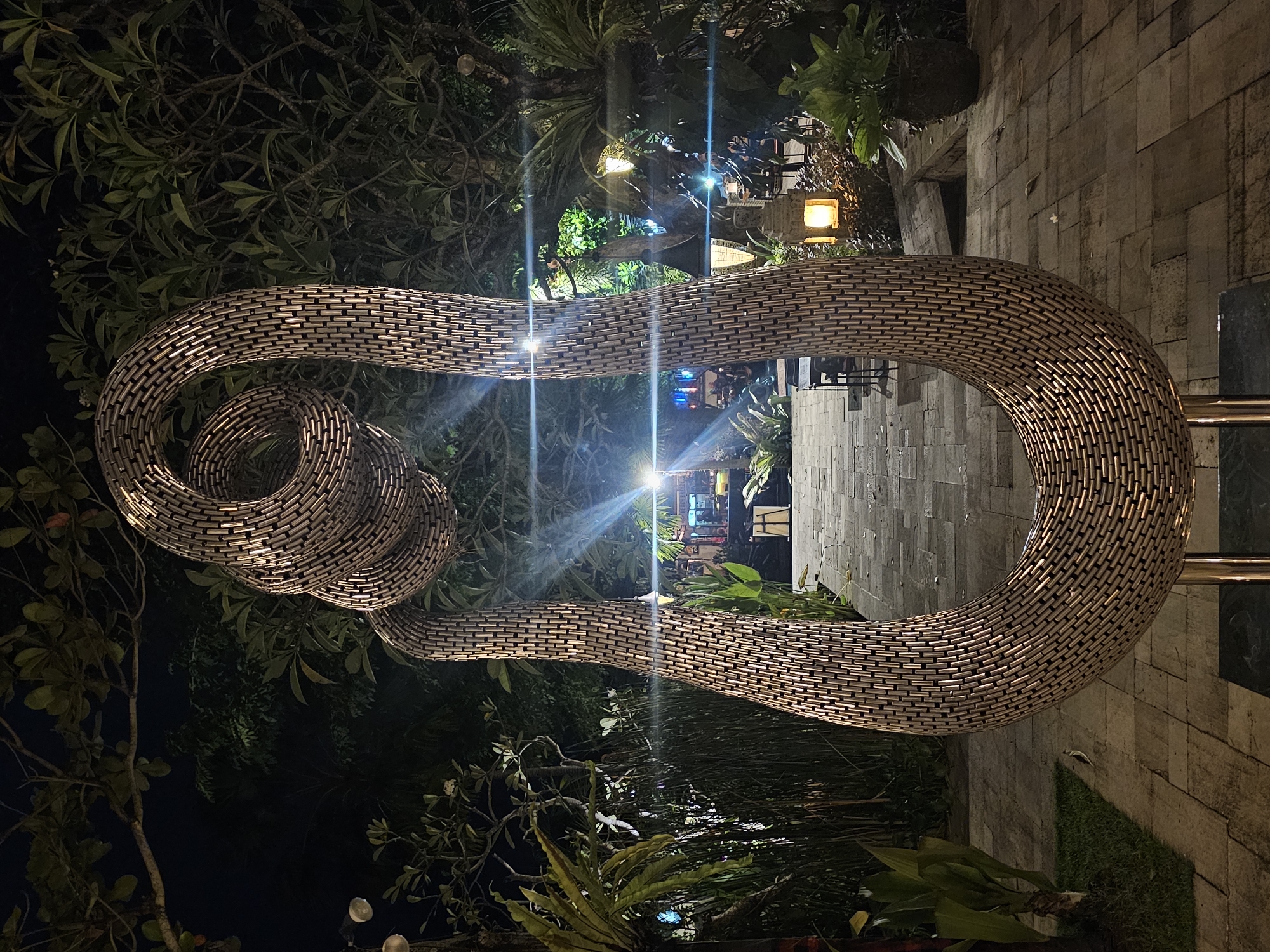 A low-angle, dimly lit, outdoor image depicts a large, curvilinear sculpture constructed of small, metallic, brick-shaped elements. The sculpture is positioned amidst dense foliage. In the background, there are indications of an urban environment. The image was taken in the city of Denpasar, Indonesia.