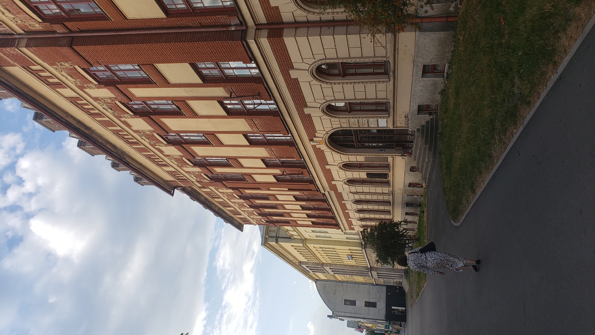 The image depicts a street scene in Prešov, Slovakia, featuring a large, multi-story building with red brick and beige facade details. The building has numerous windows with dark red frames. In the foreground, a person wearing a black and white patterned dress and carrying a black bag walks away from the camera on a paved path. The path curves to the right, bordering a grassy area. Further down the street, other buildings are visible, including one with a light yellow facade. The sky is partly cloudy. The scene appears to be a daytime exterior shot.