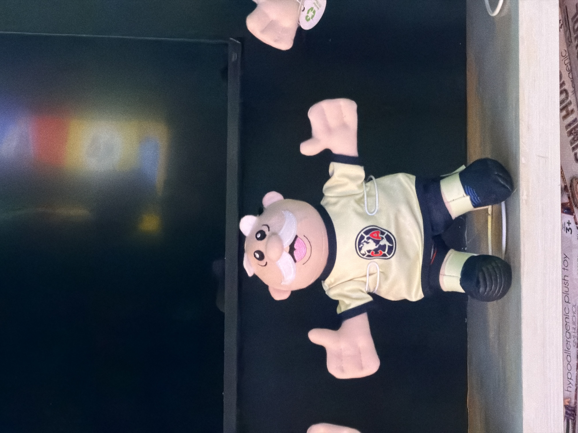 A horizontal medium shot depicts a plush toy of a cartoonish elderly man wearing a Club América soccer uniform, positioned on a shelf. The background is dark and ambiguous. Part of a black monitor is visible on the left. The box of a "hypoallergenic plush toy" is next to the shelf on the right. The specific location in Los Angeles is not discernible from the image.