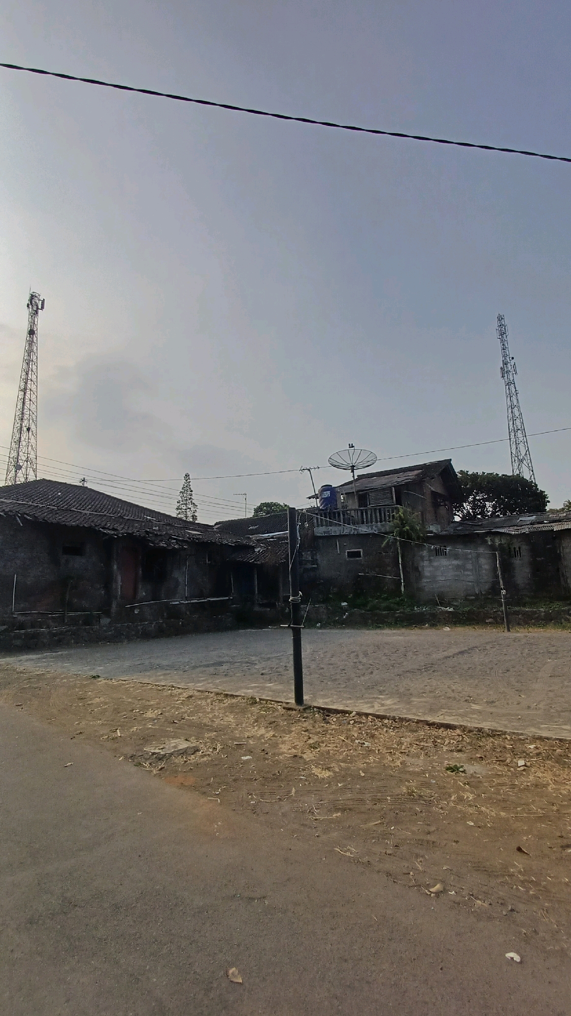 The image depicts a scene in the city of Badran, Indonesia, characterized by a slightly overcast sky and an outdoor environment. In the foreground, a paved road transitions to a dirt area with scattered debris. In the mid-ground, several buildings are visible, appearing aged with weathered roofs. A prominent feature is a satellite dish mounted on one of the buildings. Two tall telecommunication towers, typical of cell phone service infrastructure, stand in the background, one to the left and another to the right of the frame. Electrical wires are strung across the sky, connecting various points. The overall impression is that of a rural or semi-urban area with functional, albeit worn, infrastructure. There are no people or animals visible in the image, and no specific actions or interactions are occurring.