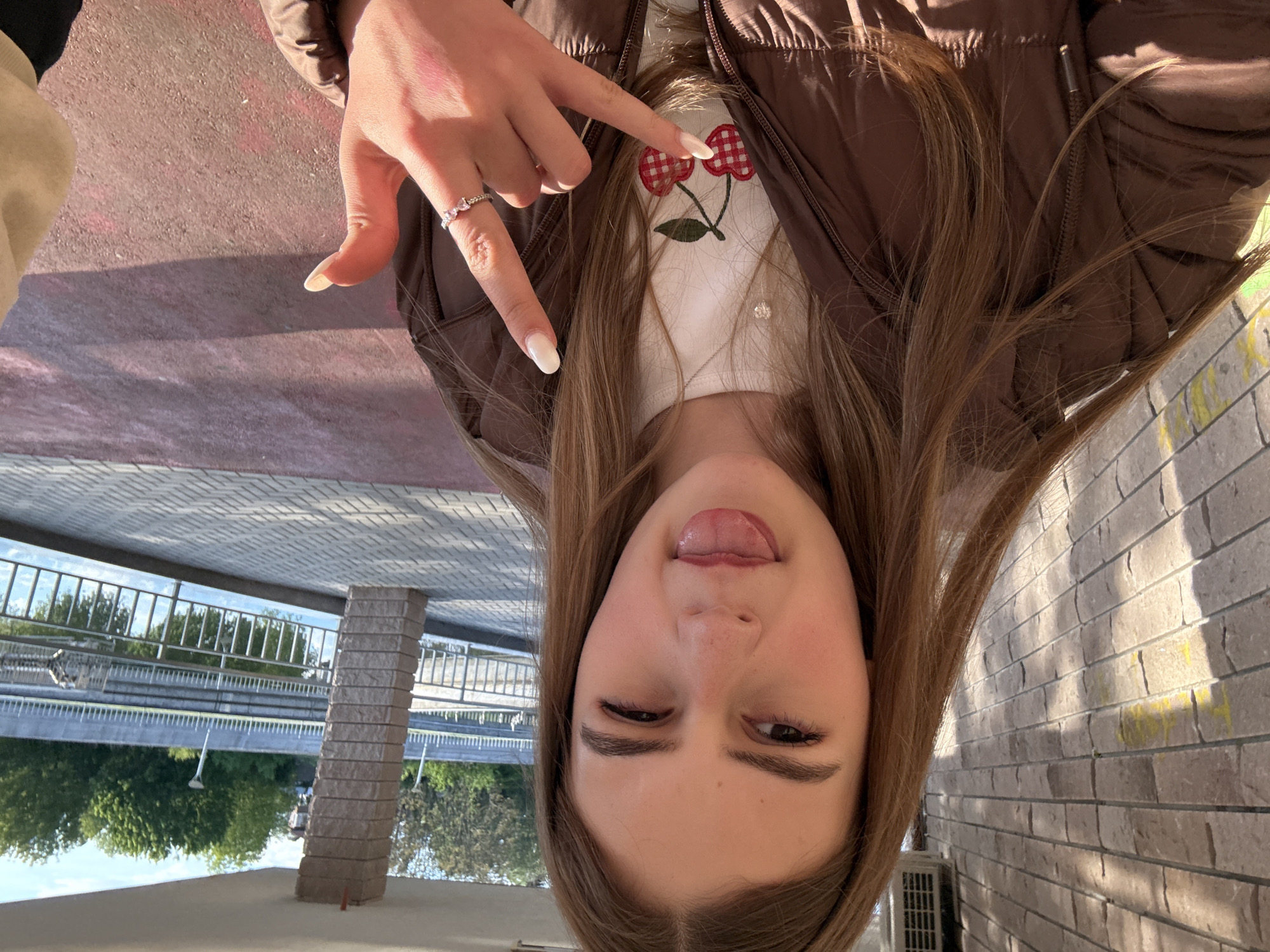 A young person is facing down towards the camera, sticking out their tongue. They are wearing a brown puffer vest over a white top with a cherry design. They have long brown hair and are making a gesture with their hand, pointing down with their index finger. A ring is visible on their finger. The background includes a brick wall and a bridge with trees in the distance. The location appears to be an urban area, possibly in Panagyurishte, Bulgaria given the prompt.