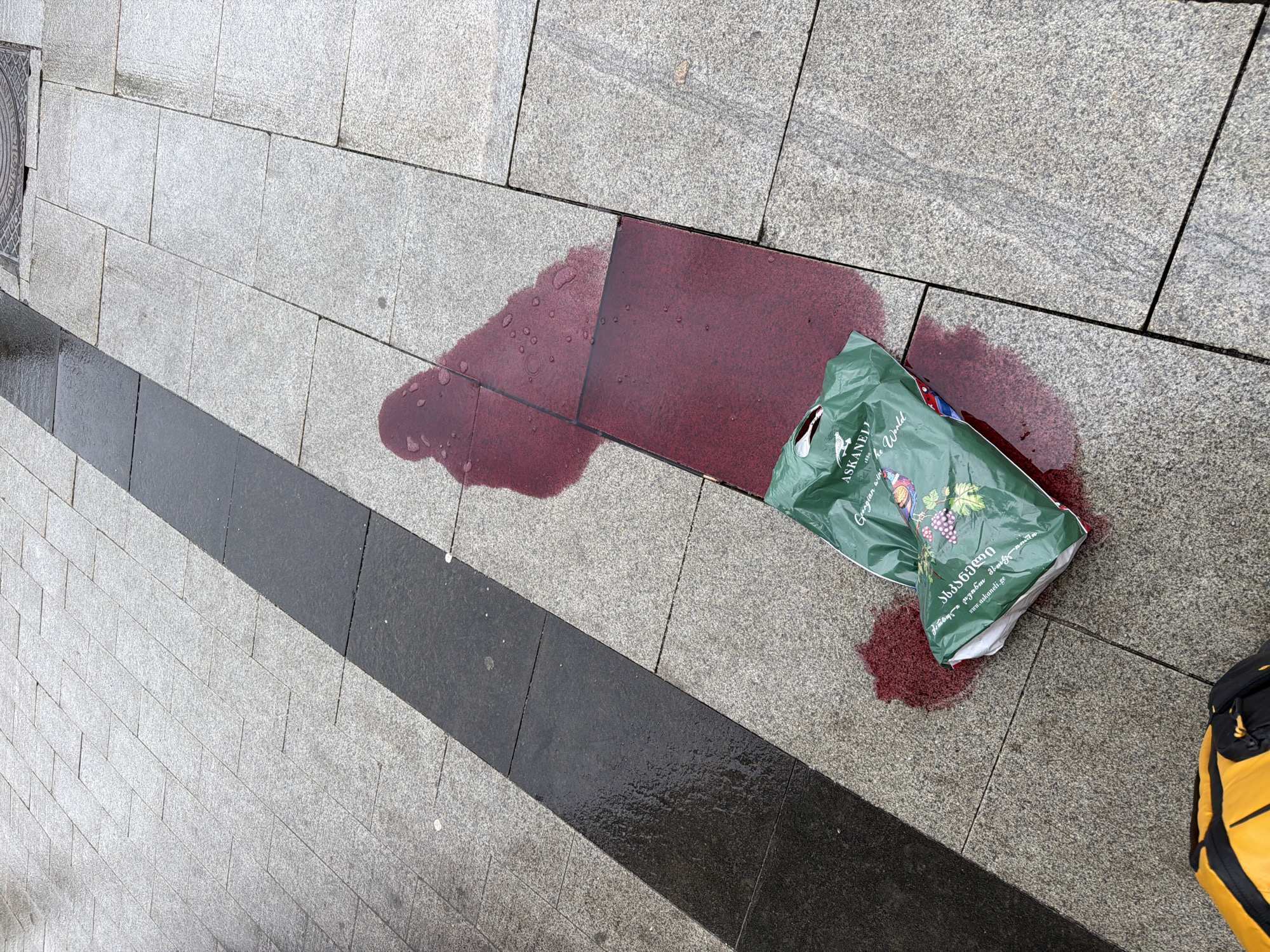 A spilled bag of red wine sits on a pedestrian walkway made of gray stone slabs. The spill has stained a red tile. The bag is green and appears to be from a shop. A black stripe runs through the stone slabs. A partial view of a yellow and black bag is visible in the lower right corner. The location is the city of Moscow, Russia.