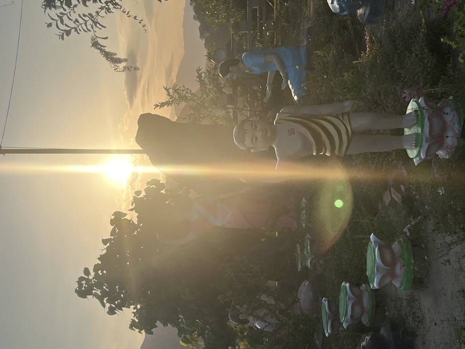 The image depicts an outdoor garden scene at sunrise or sunset in Nha Trang, Vietnam, marked by significant backlighting and a prominent vertical sun flare originating from the upper-center. In the foreground, several lotus-shaped pedestals with light green tops and white-pink bases are arranged on a gently sloped, vegetated ground. A central, light-skinned statue of a young male figure stands on a similar lotus pedestal; he wears a yellow and brown striped cloth draped over his left shoulder and torso, exposing his right shoulder and legs, with a red swastika symbol on his chest. His right arm is extended outwards, and his left arm is positioned alongside his body.
In the midground, to the left of the central figure, a larger, pink-skinned statue of a standing figure is partially obscured by dense green foliage and the sun's glare. To the right of the central figure, a blue-skinned female statue kneels or sits, with both hands extended forward. A dark, irregularly shaped rock formation or statue is silhouetted behind the central figure. The background reveals hazy distant mountains under a light orange sky with scattered clouds. A tall, thin vertical pole is visible to the right, near the sun's direct glare.