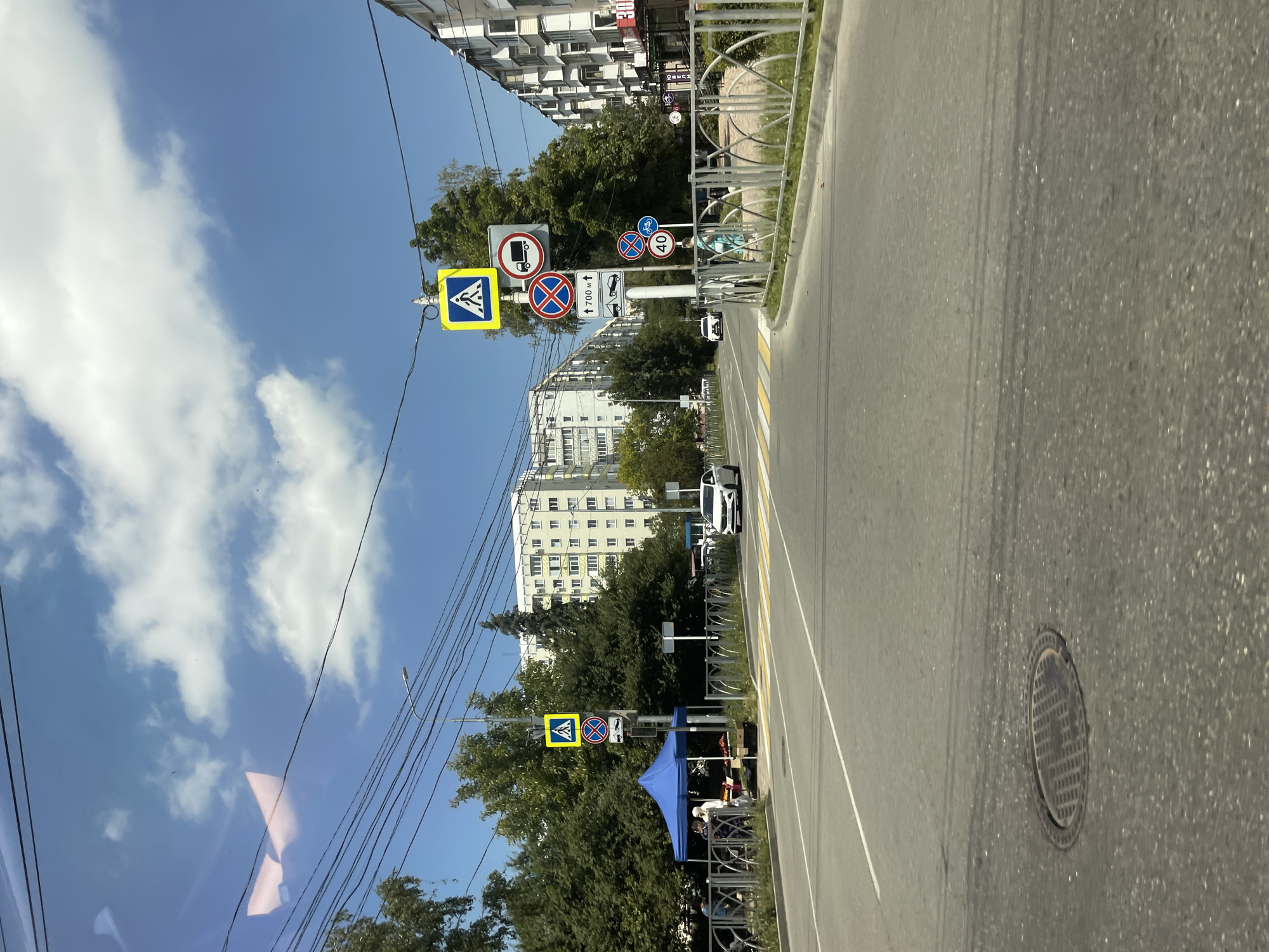 The image captures a street scene in Stavropol, Russia, on a clear day with scattered clouds. A road runs through the center of the frame, marked with white lines and a pedestrian crossing. Traffic signs are visible, including a pedestrian crossing sign, a truck restriction sign, a "no stopping" sign, and a speed limit sign indicating 40 km/h. Several vehicles are present on the road, including a white car and a glimpse of another vehicle. On the left side of the road, a blue tent is set up, possibly for a market or event, with several people gathered around it. Trees line the street, and apartment buildings are visible in the background. A metal fence runs along the side of the road. The overall impression is that of a typical urban environment on a sunny day.