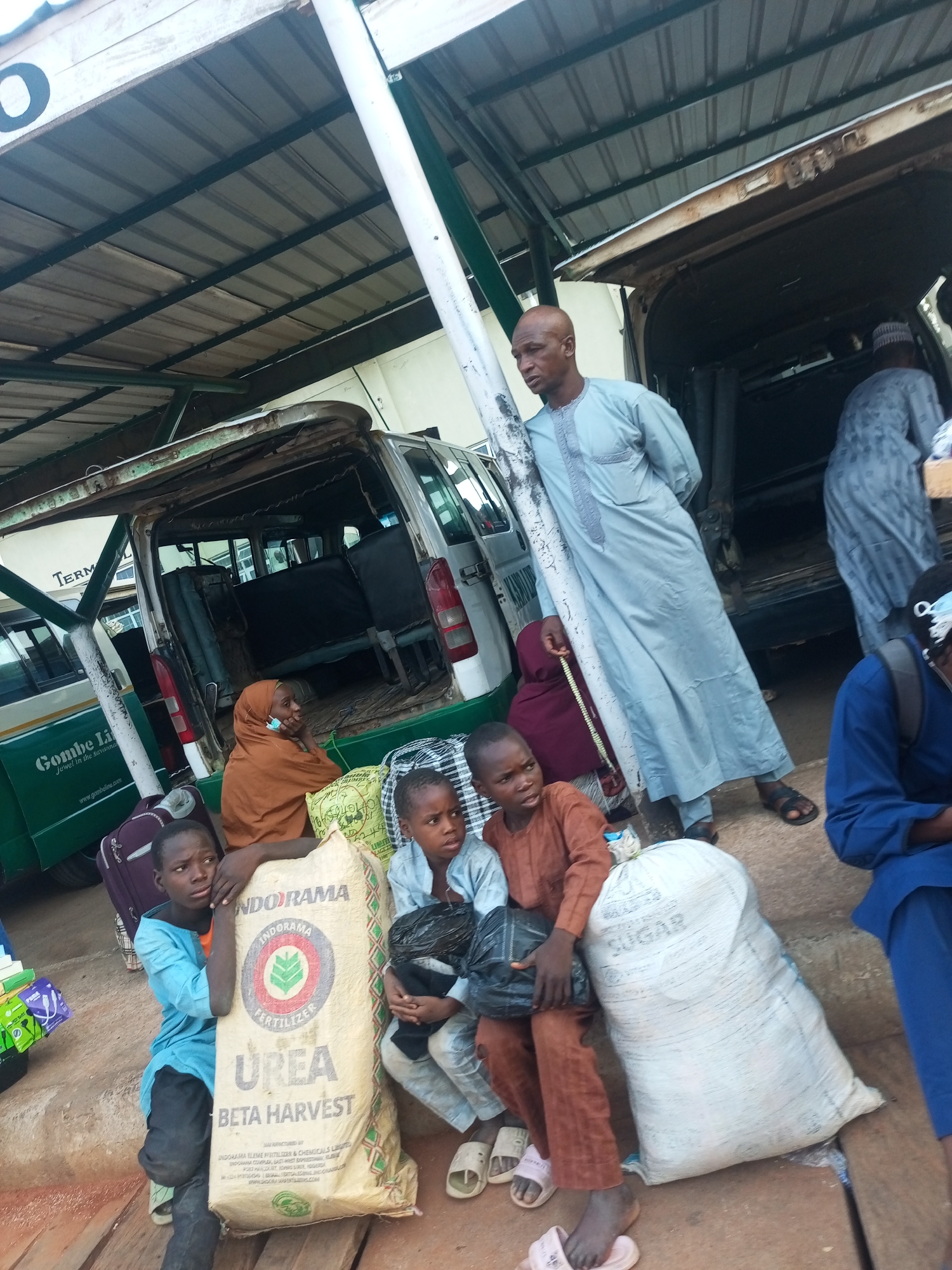 The image depicts a scene at a transport terminal in Gombe, Nigeria. Several people, including children, are waiting with their belongings. A middle-aged man in a light blue robe stands near the open back of a van. Some of the travelers are sitting on or next to large sacks labeled "UREA BETA HARVEST" and "SUGAR." Luggage and other items are scattered around the waiting area. A bus with "Gombe Line" printed on the side is visible. The scene appears to be a typical transportation hub with travelers and their goods.