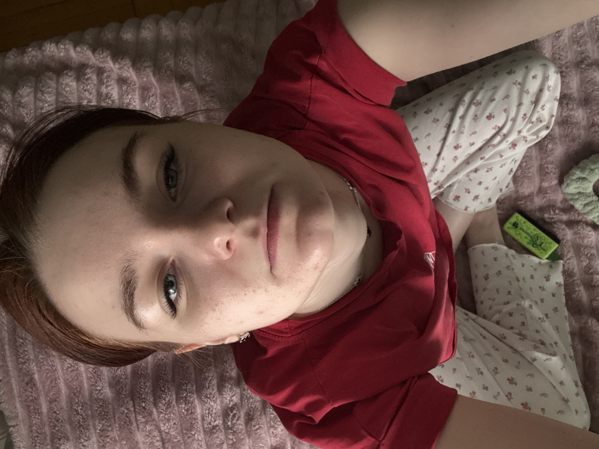 A high-angle shot shows a woman in a red t-shirt and floral pajama pants. She is sitting on a pink, textured blanket, looking upwards at the camera. A green and yellow object and a green knotted object are visible near her legs. The location is unknown.