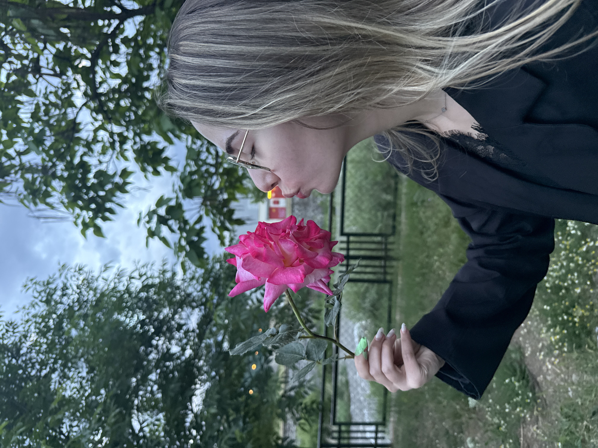 A woman is positioned outdoors, likely in a garden or park, with trees and foliage in the background, possibly in Blagoevgrad, Bulgaria. She is holding a large pink rose close to her face, seemingly about to smell or kiss it. The woman has long blonde hair, wears glasses, and is dressed in a black jacket or blouse. The background includes hints of a fence and other structures. Her nails are painted with a light-colored polish and a small green dot on each nail. The orientation of the image is such that the woman appears to be positioned horizontally.