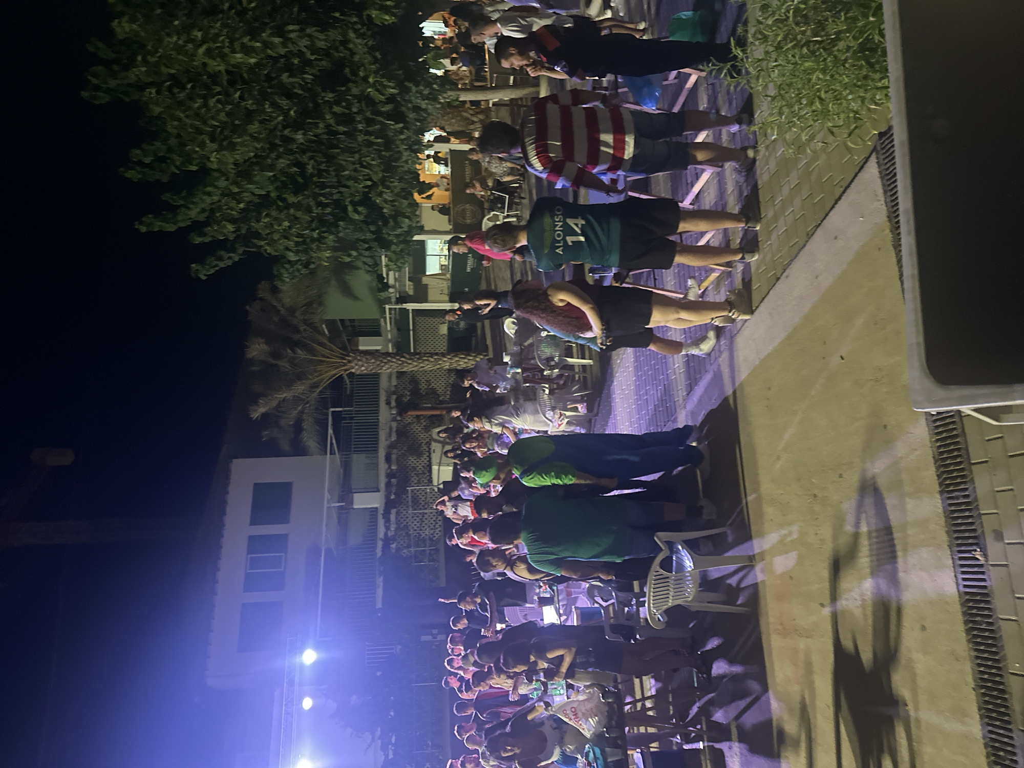 The image depicts an outdoor nighttime gathering in Blanca, Spain. A crowd of people are standing around tables and chairs, some holding drinks, suggesting a social event or party. A stage with bright lights is visible on the left side of the frame, indicating a possible performance or DJ set. The background shows buildings with windows and balconies, and trees, including a palm tree, are present in the scene, suggesting a warm climate. A crane is also visible in the distance, hinting at ongoing construction or development. The overall atmosphere appears lively and casual.