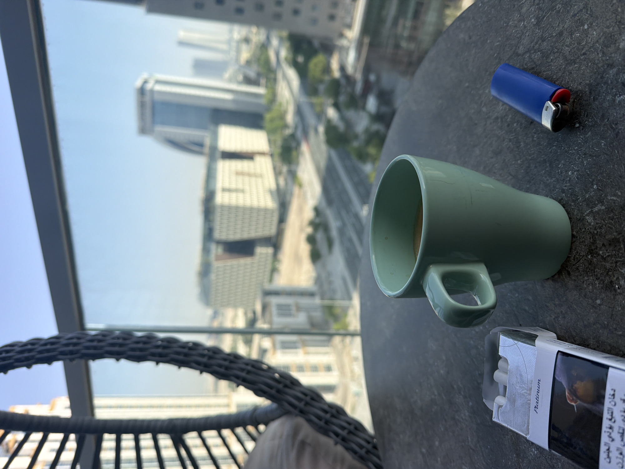 A pack of "Platinum" cigarettes and a blue lighter rest on a dark, textured table beside a light green mug of coffee. The mug is partially visible, with the coffee's surface reflecting the light. In the background, a blurred cityscape unfolds through a large window. Tall, modern buildings with distinct architectural features dominate the view, interspersed with roads and some greenery. The sky is a clear, bright blue, suggesting daytime. A portion of a woven chair is visible in the foreground on the left.