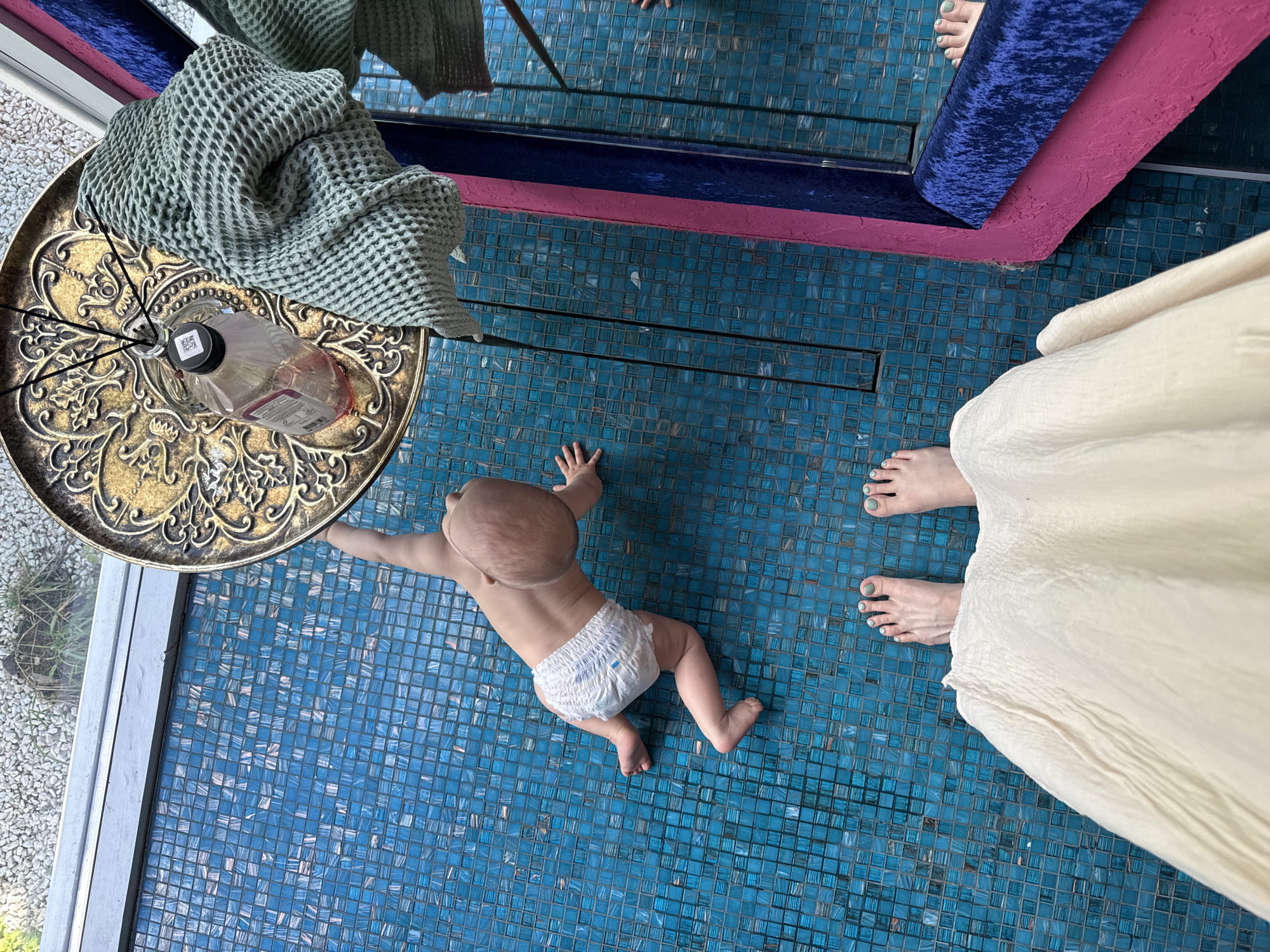 A baby in a diaper is crawling on a blue-tiled floor. A person's feet and white dress are visible near the baby. An ornate gold-colored table with a green towel and diffuser sits to the left of the baby. A mirror with a blue and pink frame is in the upper portion of the image. The scene is indoors. There is no visual information to support this scene being in Чёрное, Russia.