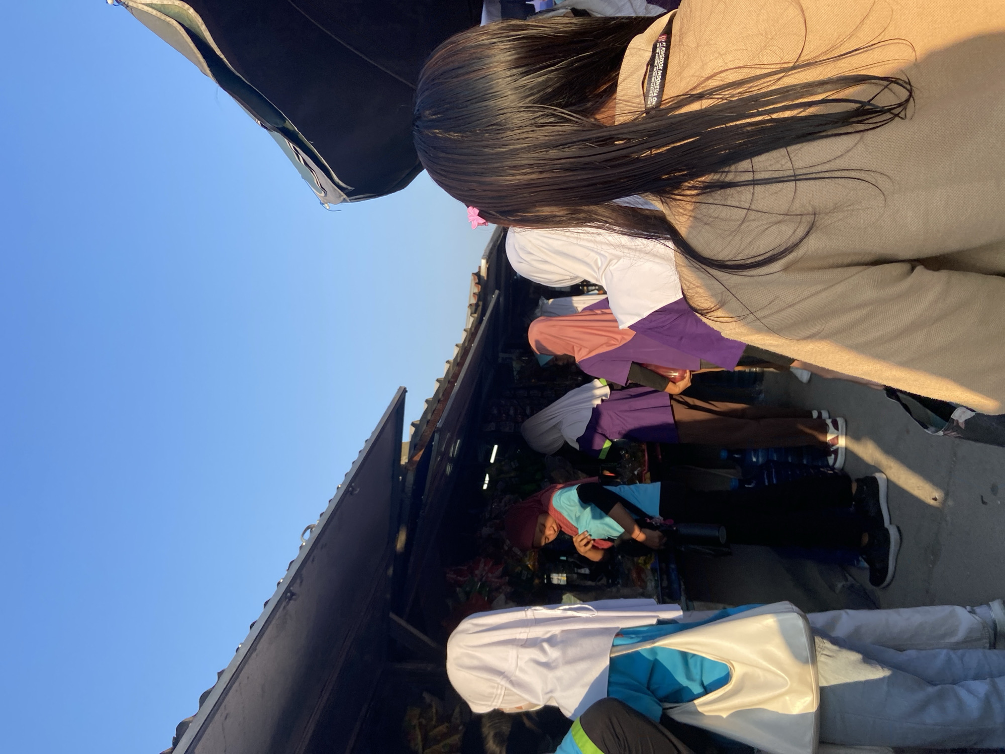 In the city of Tanjungrejo, Indonesia, the image captures a street scene with several individuals standing by market stalls. The time of day appears to be late afternoon, indicated by the warm sunlight casting long shadows on the ground. The sky is a clear, bright blue, suggesting good weather.
In the foreground, a person with long, dark hair, adorned with a small pink flower, is seen from behind, wearing a light brown jacket with a visible tag that reads "FT JUNGKOOK INDONESIA".
Further into the scene, a group of people are gathered around market stalls that are covered by dark awnings. Several individuals are wearing hijabs in various colors, including white, purple, and pink. One person in a turquoise shirt and black pants is looking down at something on the stall. Another person in a purple top and brown pants is also visible. The stalls appear to be selling various goods, with bags and bottles visible. The ground is paved and appears to be a public walkway or street. The overall atmosphere suggests a bustling market or shopping area.